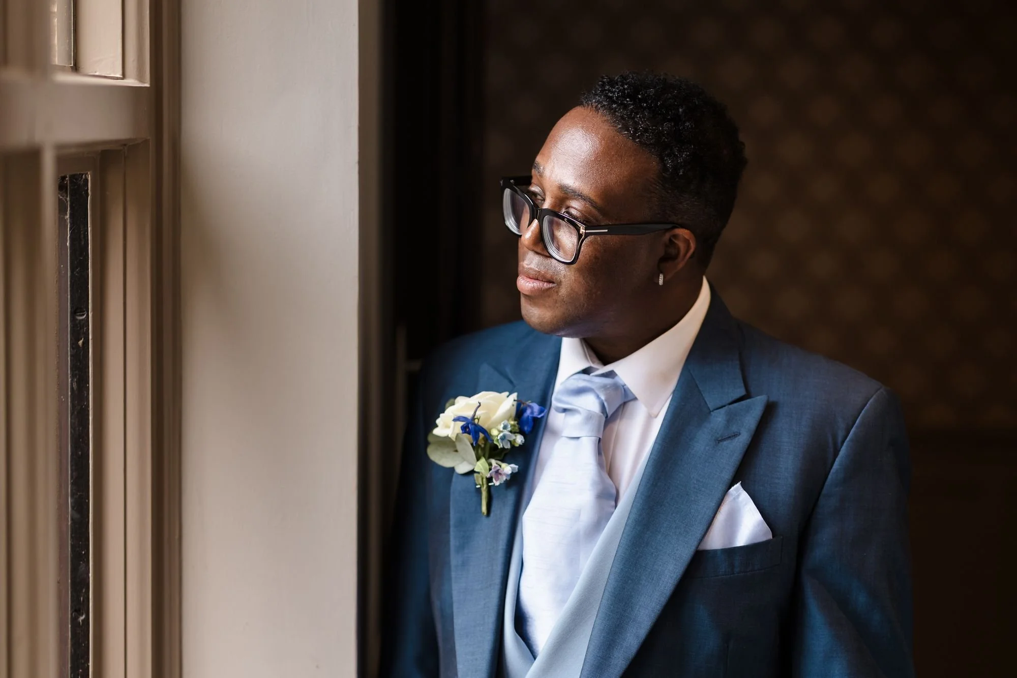 One of the grooms in a blue suit and glasses looking out of a window in soft natural light before the ceremony