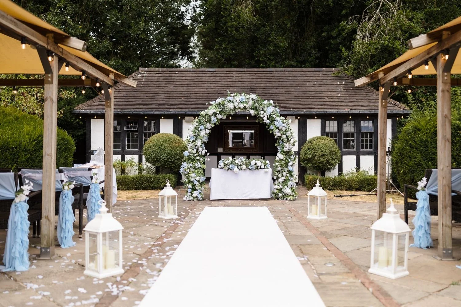 White aisle runner lined with lanterns and pale blue chair sashes leading to the floral arch at the Pump Rooms ceremony area