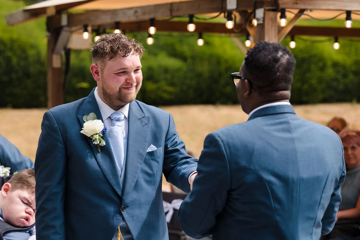 One of the grooms smiling at his partner as they face each other under the canopy during the outdoor ceremony