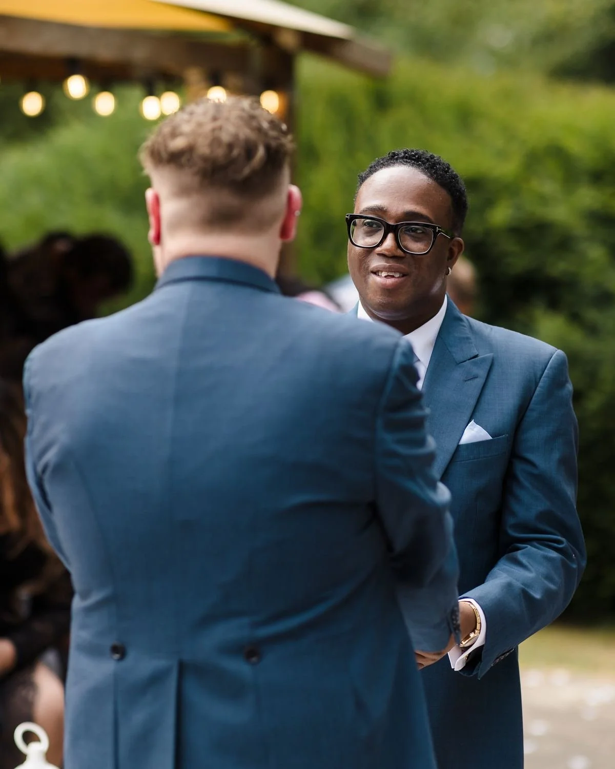 One of the grooms in glasses smiling as he holds his partner's hands during the vows, with festoon lights behind