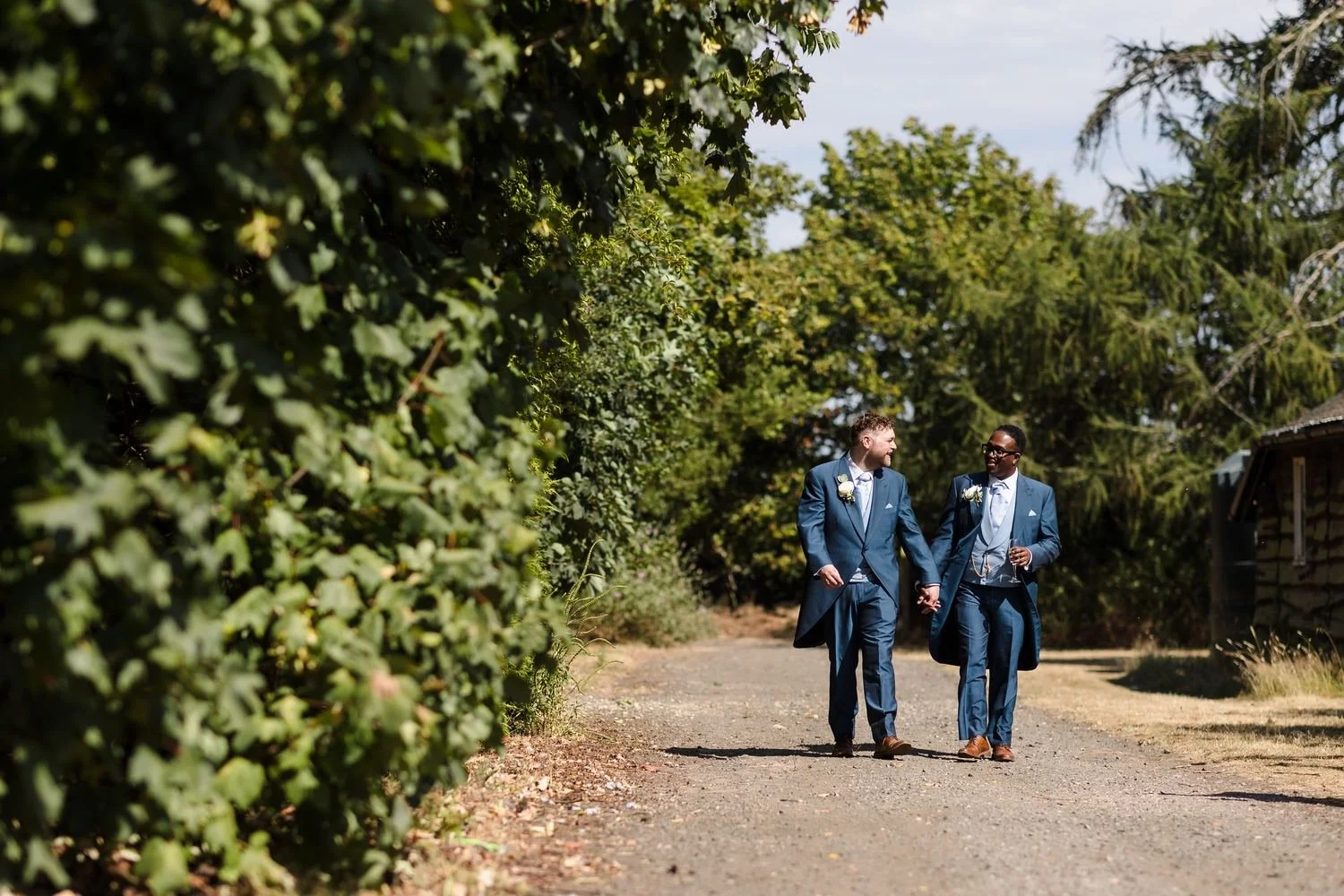 Liam and Shaun laughing as they walk hand in hand along a sunny tree-lined track in the grounds