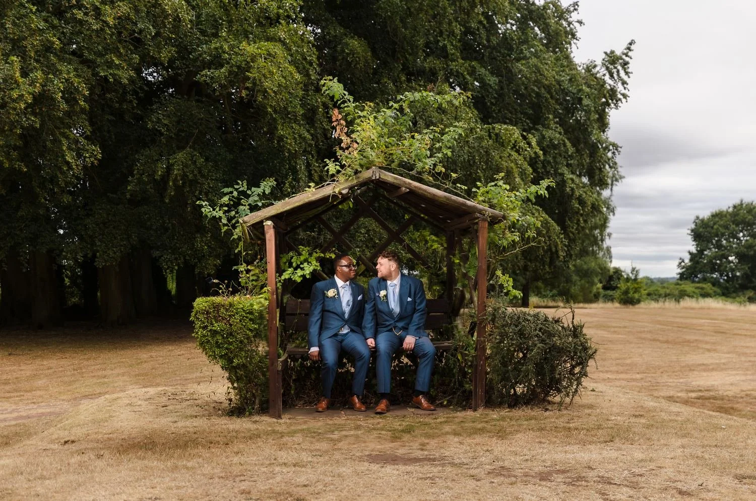 The grooms sitting together in a wooden arbour seat on the edge of the lawn, framed by mature trees