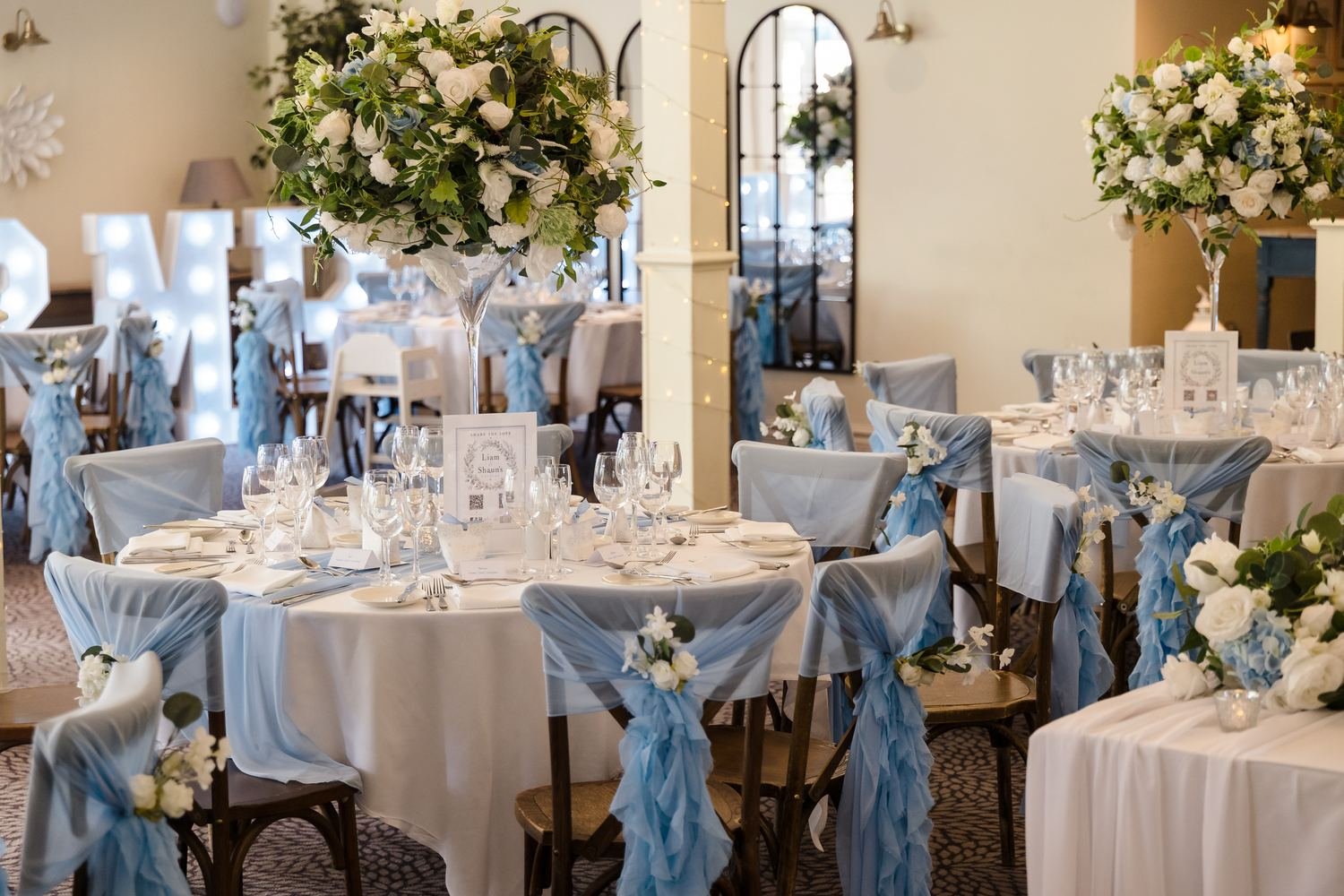 Garden Room tables laid for the wedding breakfast with white linen, pale blue chair sashes and tall floral centrepieces
