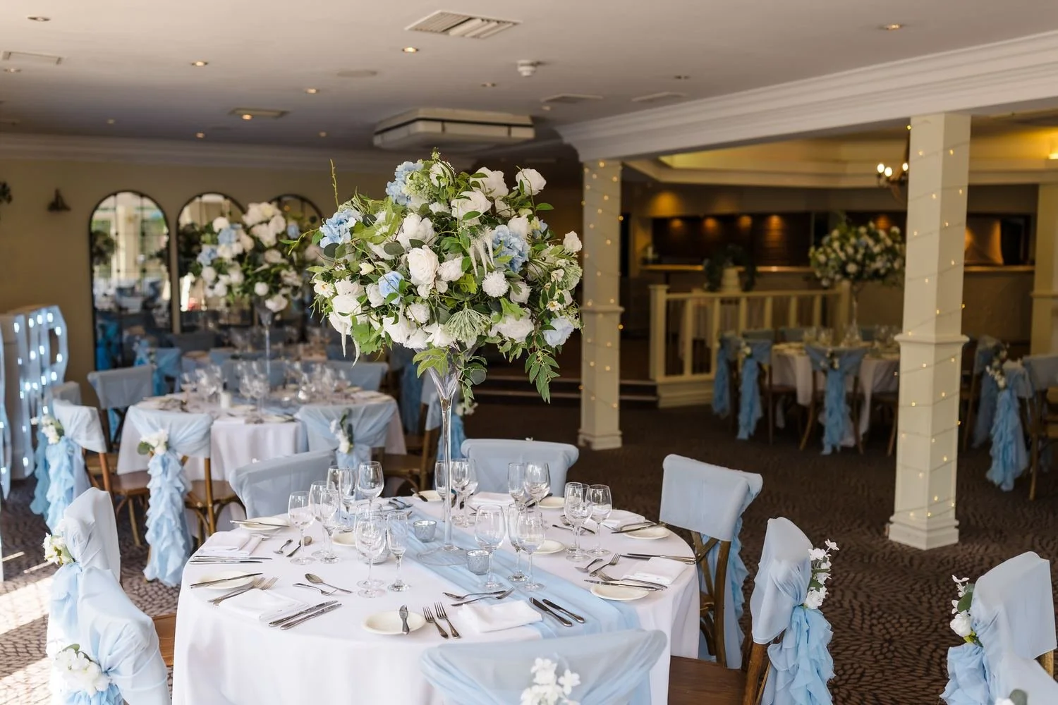 The Garden Room at Hogarths Stone Manor styled in white and pale blue, with fairy-lit columns and large flower arrangements