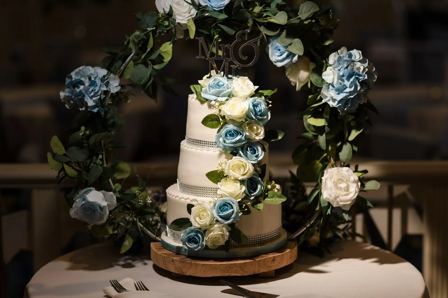 Three-tier white wedding cake with cascading blue and white roses and a Mr and Mr topper, framed by a floral hoop
