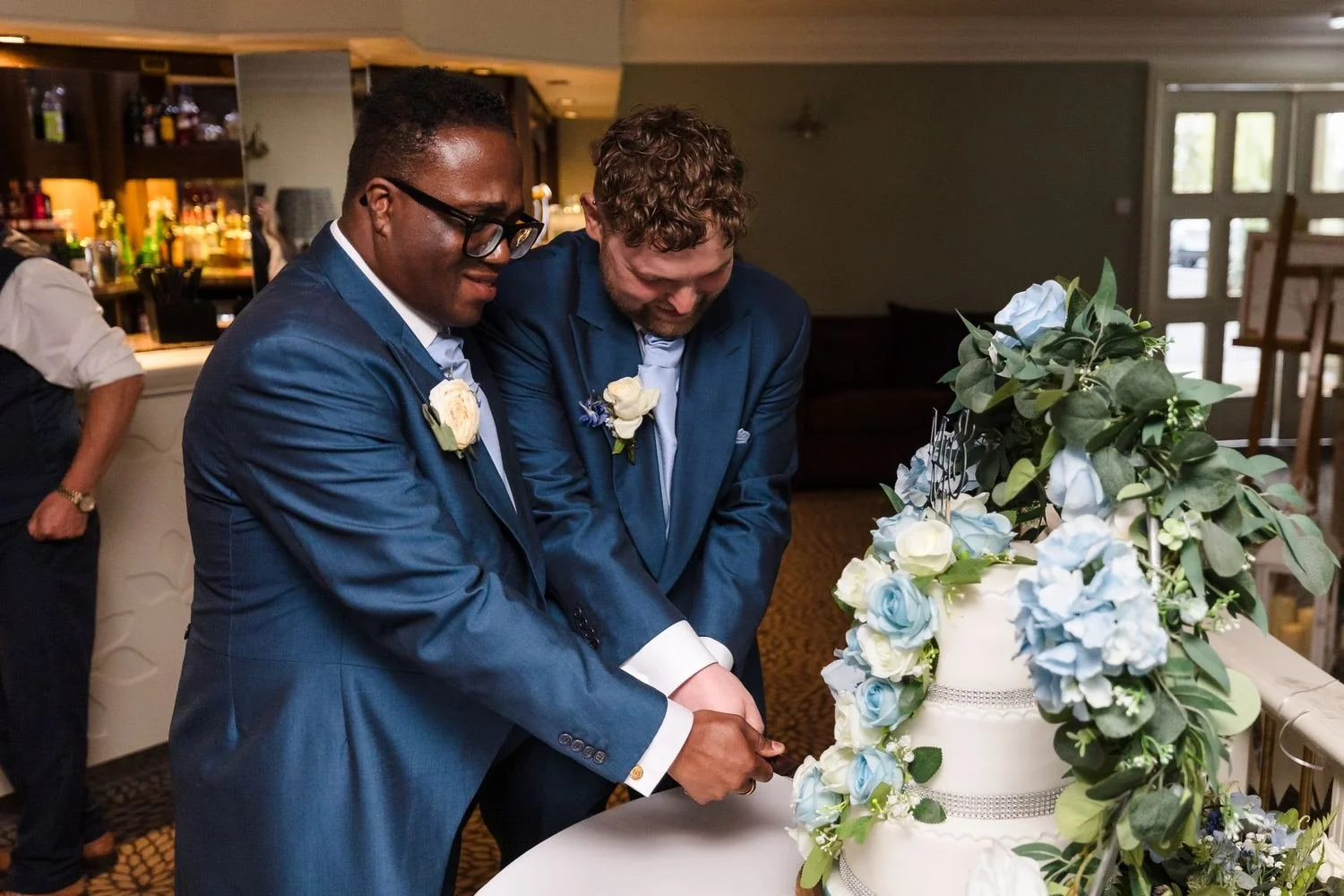 Liam and Shaun smiling as they cut their blue and white floral wedding cake together