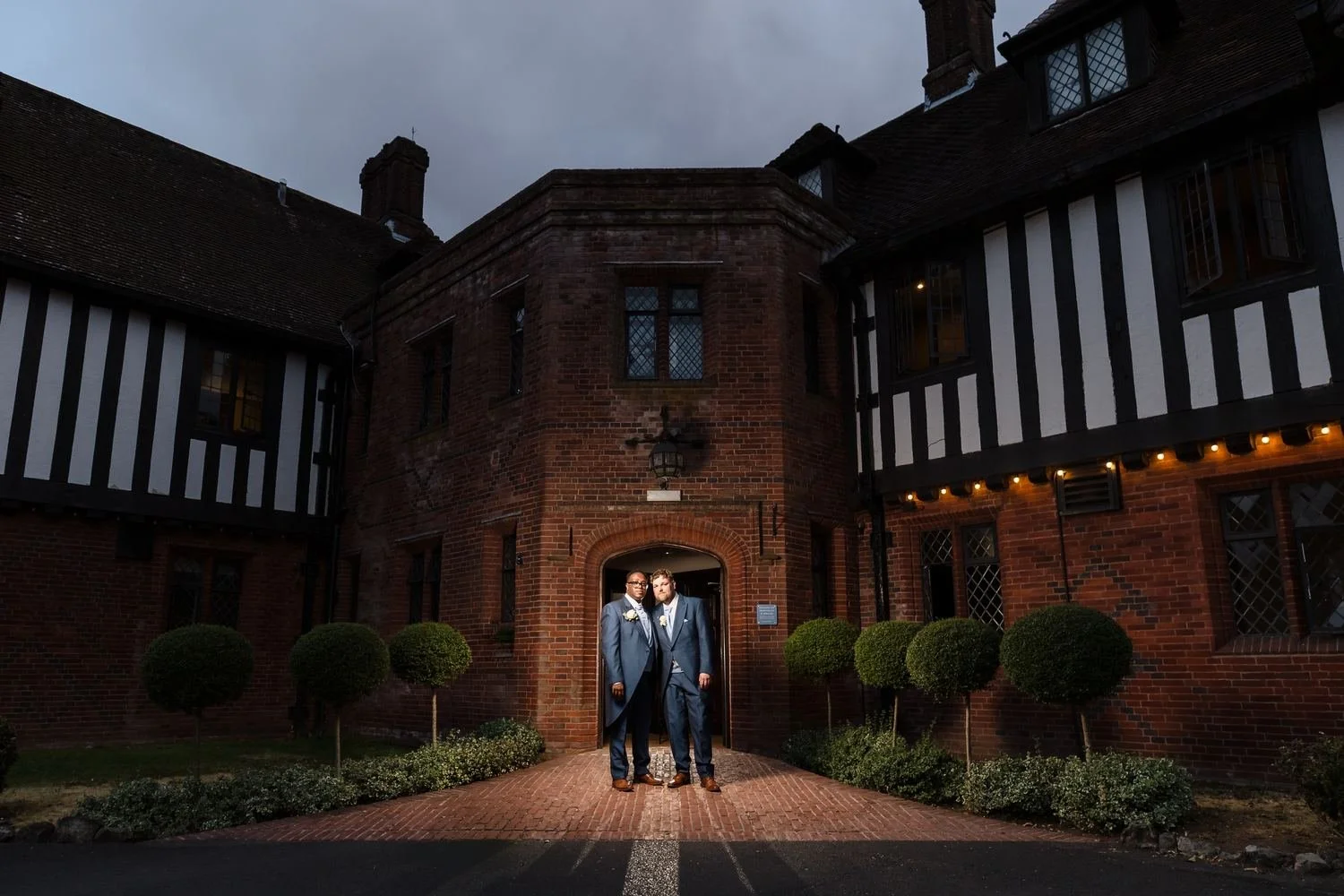 Liam and Shaun standing in the arched brick doorway of Hogarths Stone Manor at dusk, lit against the timbered building