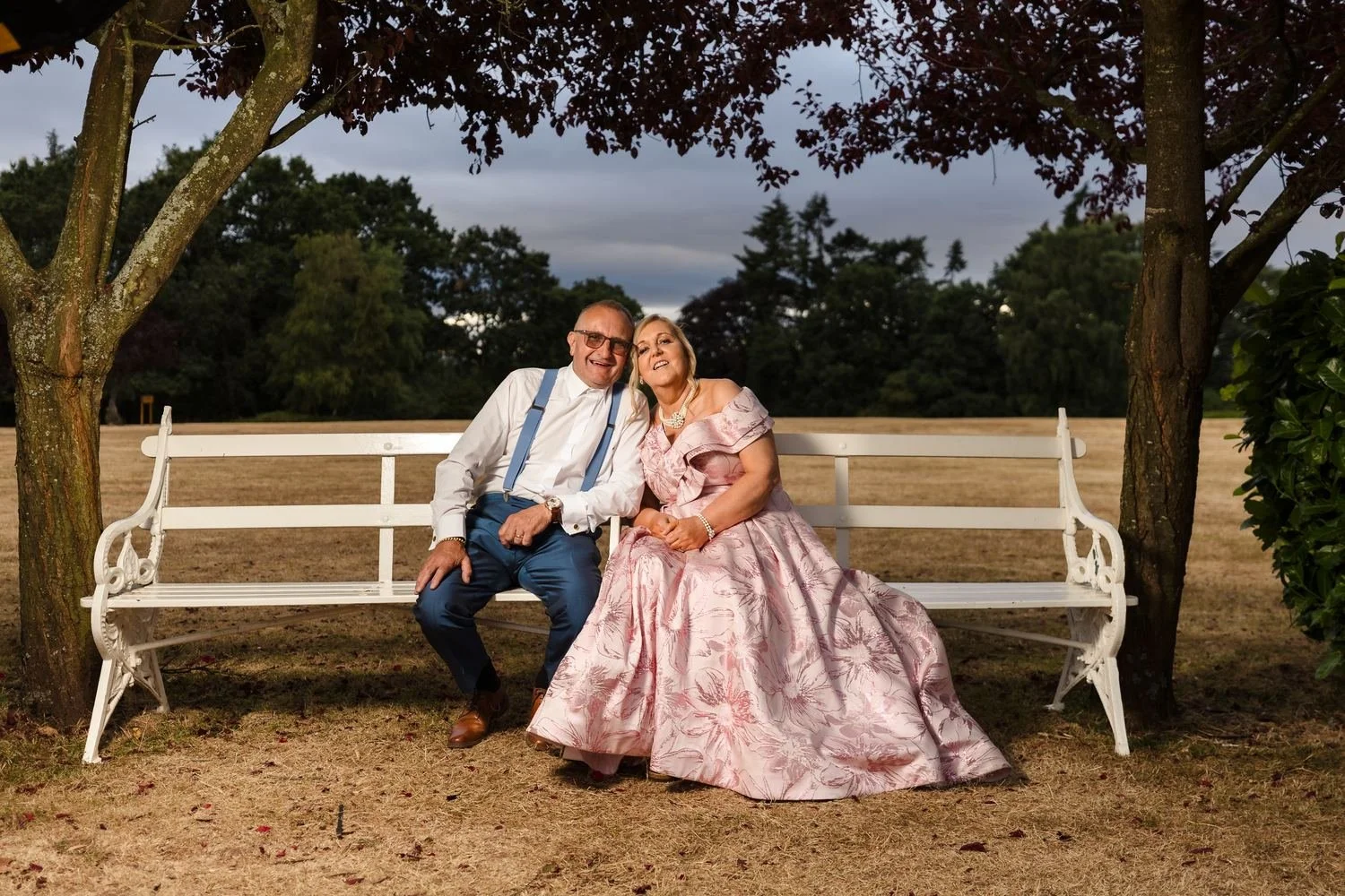 Two wedding guests sitting together on the white bench beneath the red-leaved trees in the evening light