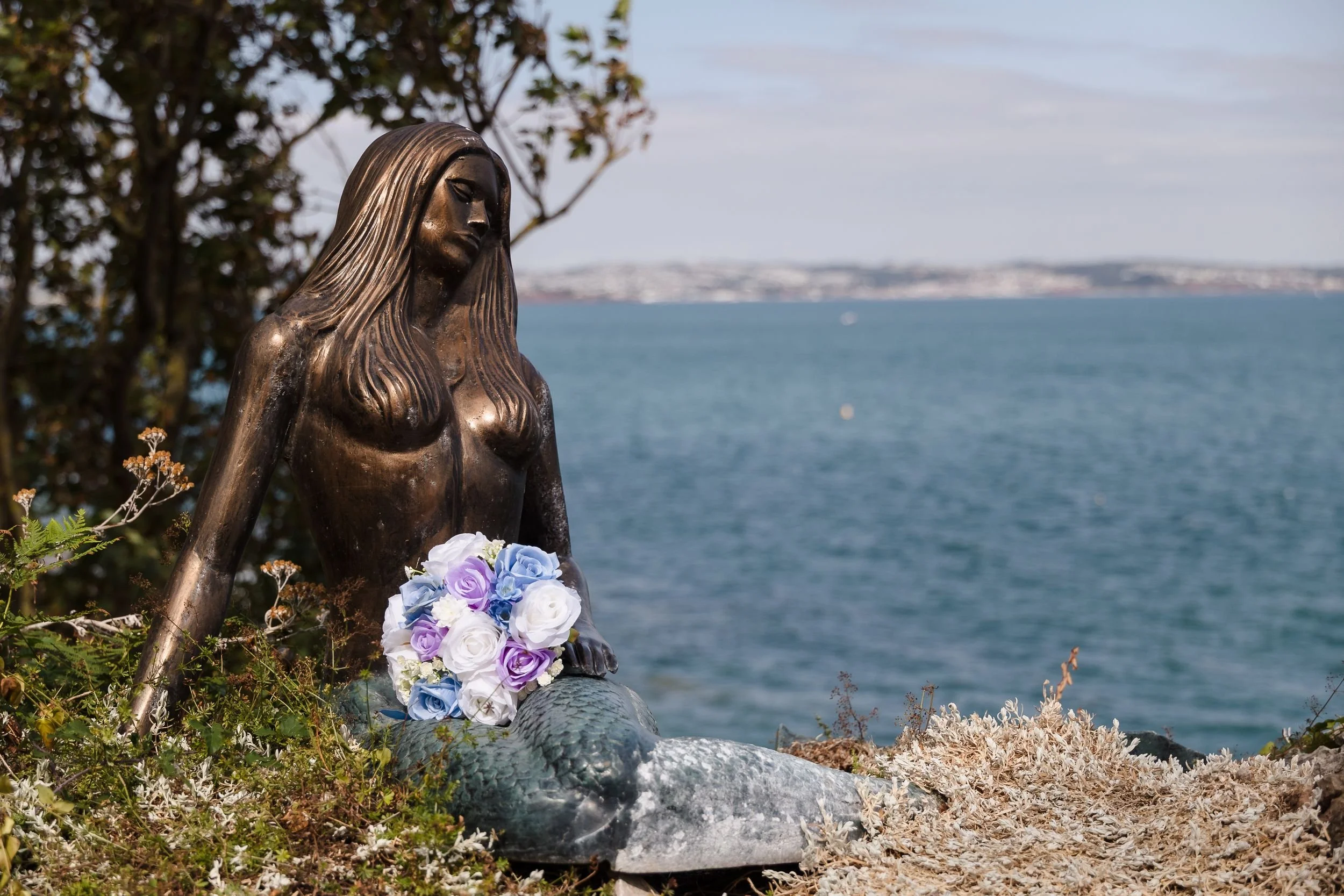 Blue, lilac and white bridal bouquet resting on the mermaid statue at Berry Head Hotel, overlooking the bay