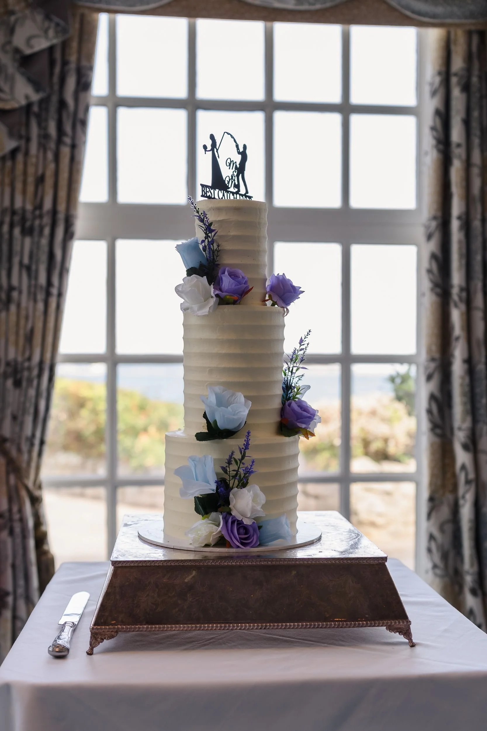 Three-tier white wedding cake with blue and lilac flowers in front of a sea-view window at Berry Head Hotel