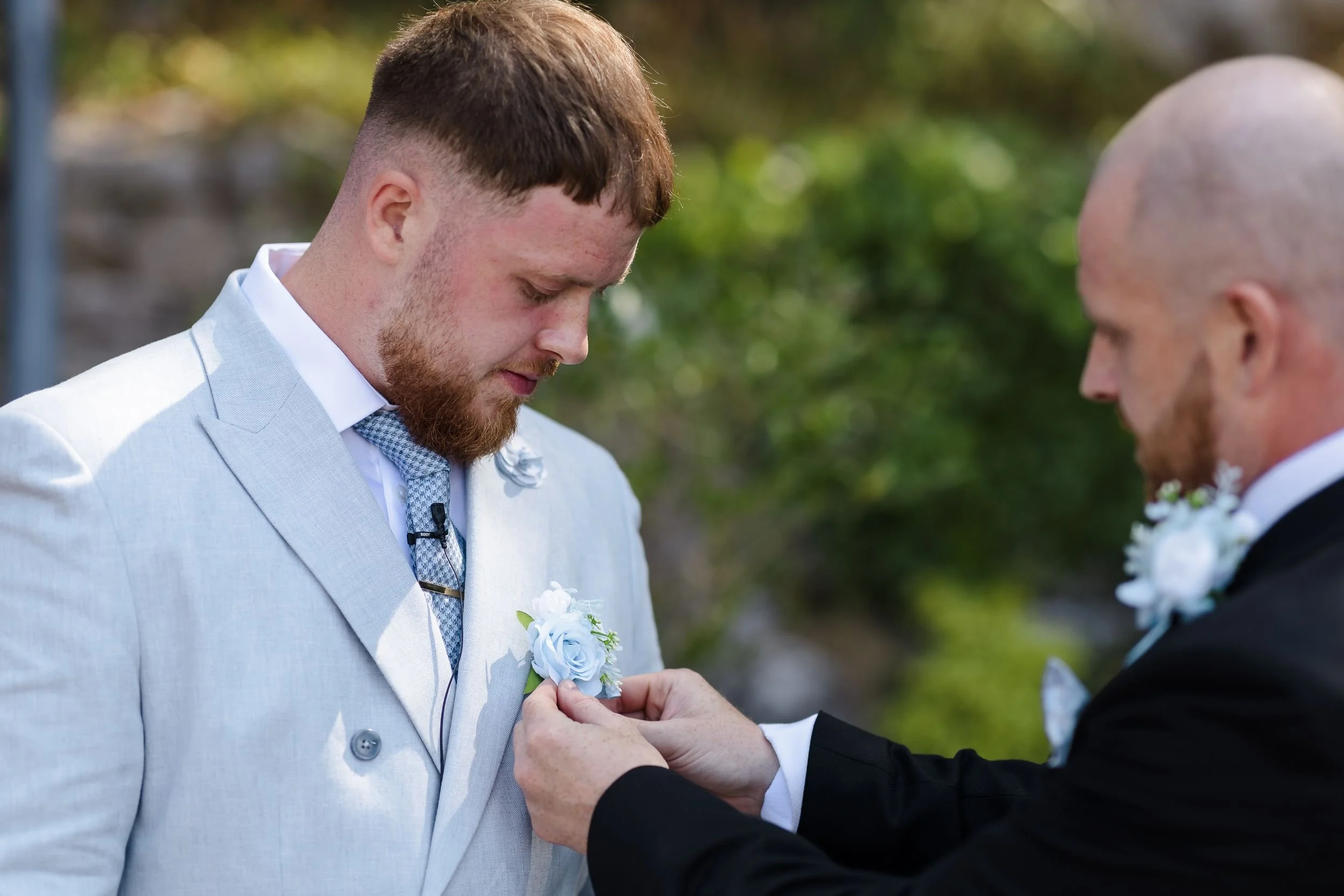 Best man pins a pale blue buttonhole to the groom's jacket before the outdoor ceremony at Berry Head Hotel
