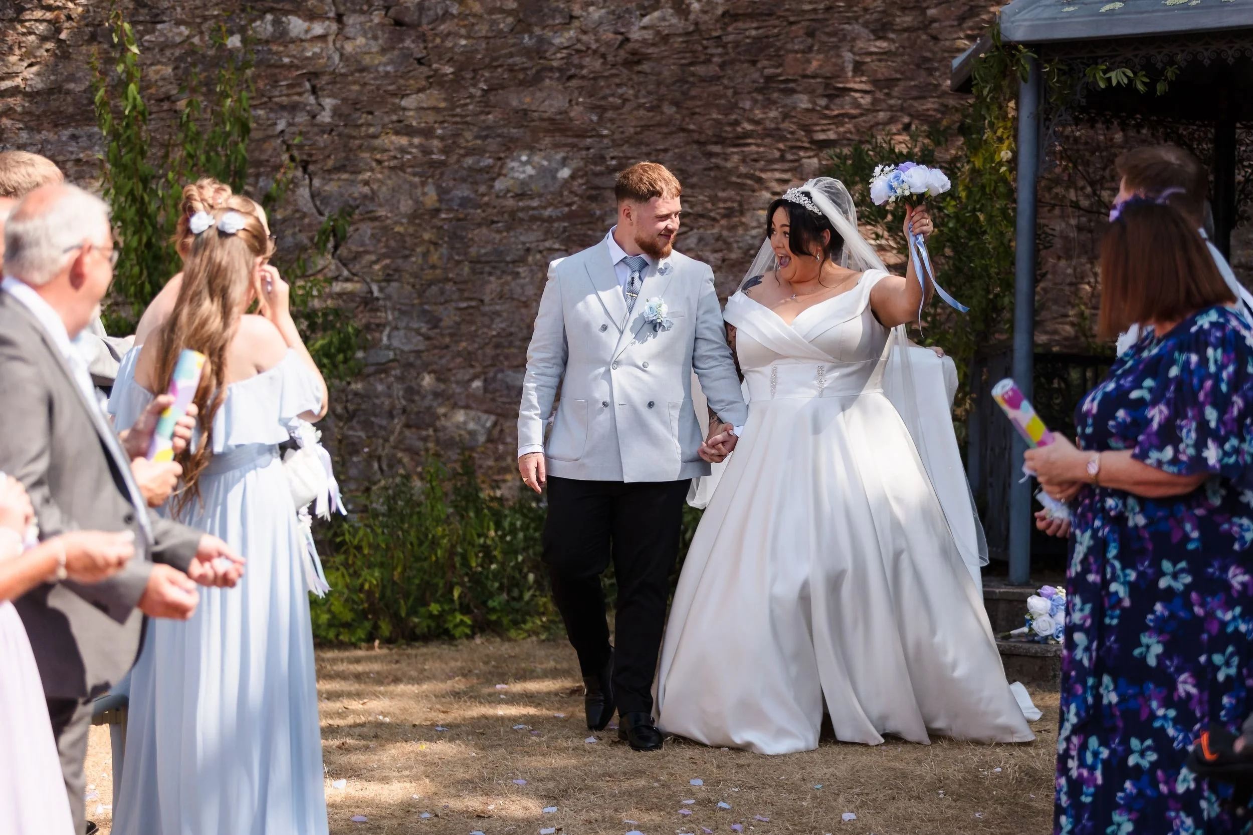 Newly married couple walk back up the aisle hand in hand, the bride raising her bouquet, at Berry Head Hotel