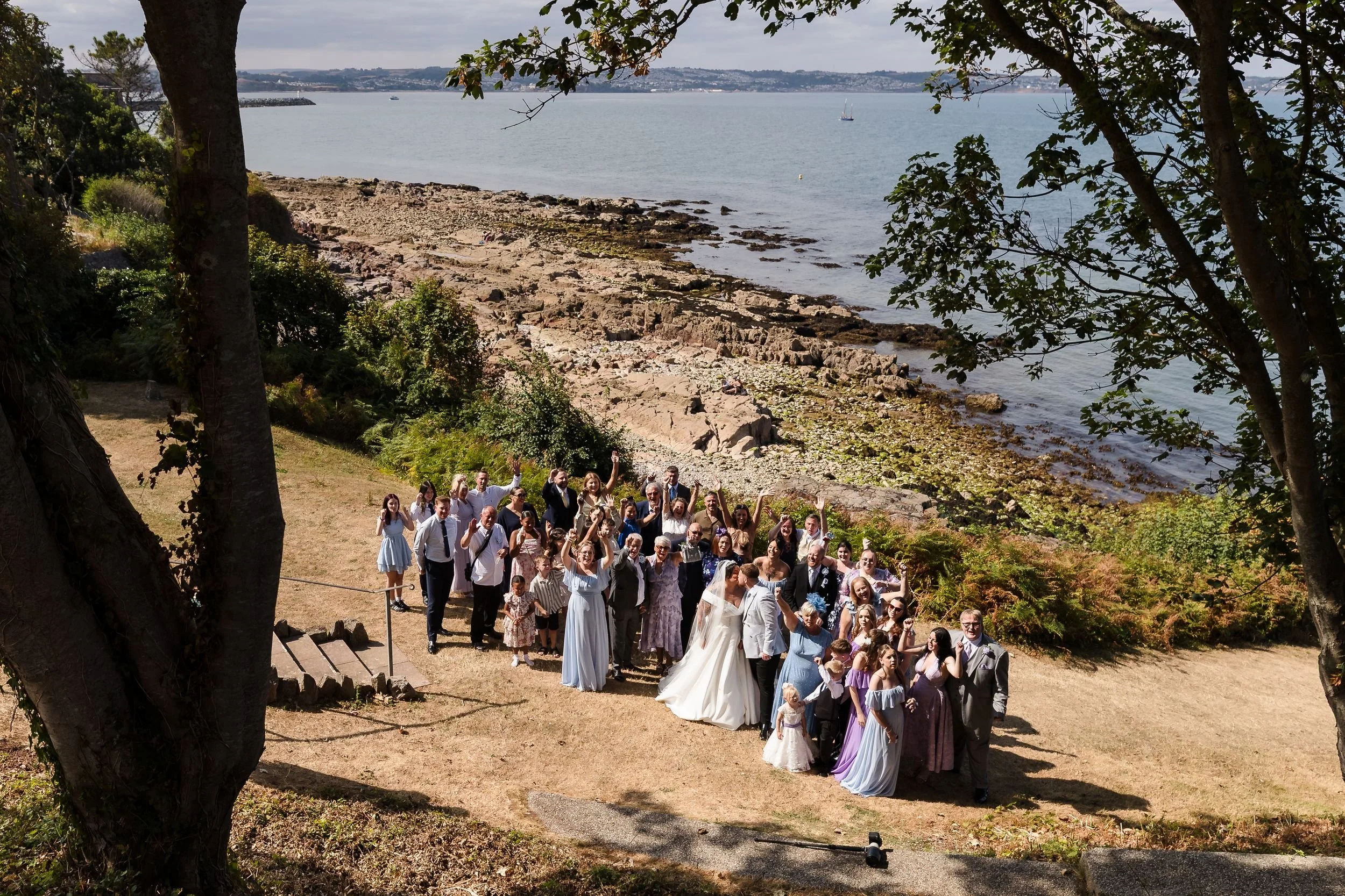 Full wedding group cheering on the lawn at Berry Head Hotel, photographed from above with the rocks and bay behind