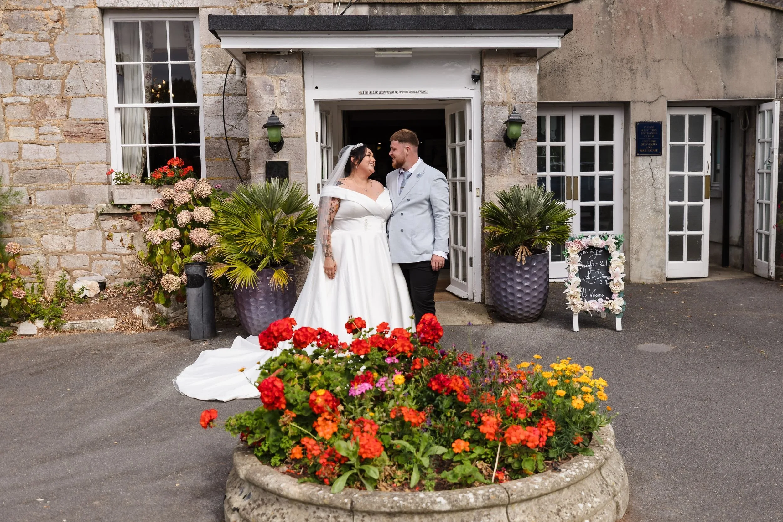 Bride and groom outside the stone entrance of Berry Head Hotel in Brixham, behind a bed of red and orange flowers