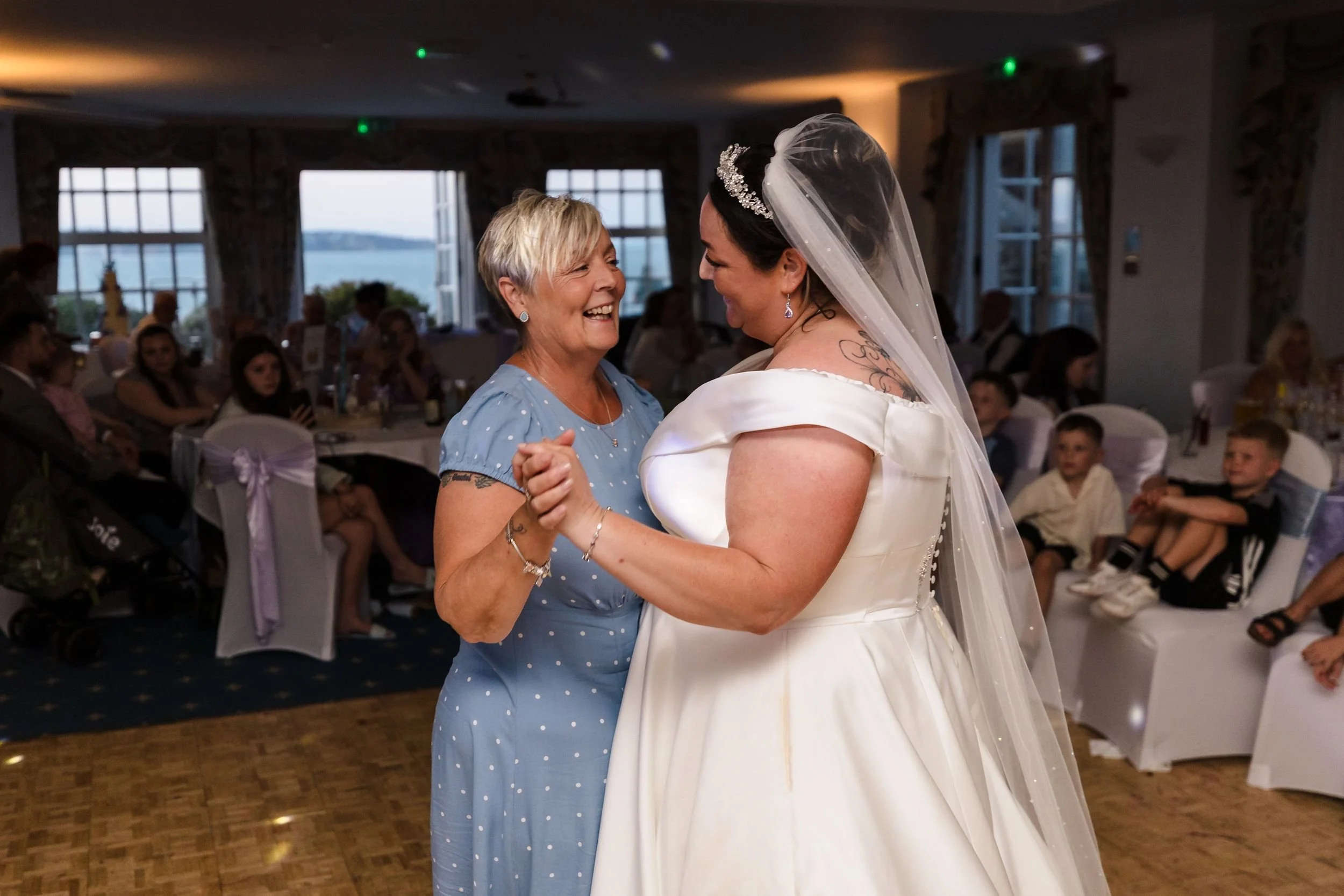 Bride dances with a family member at her evening reception at Berry Head Hotel, the bay visible through the windows