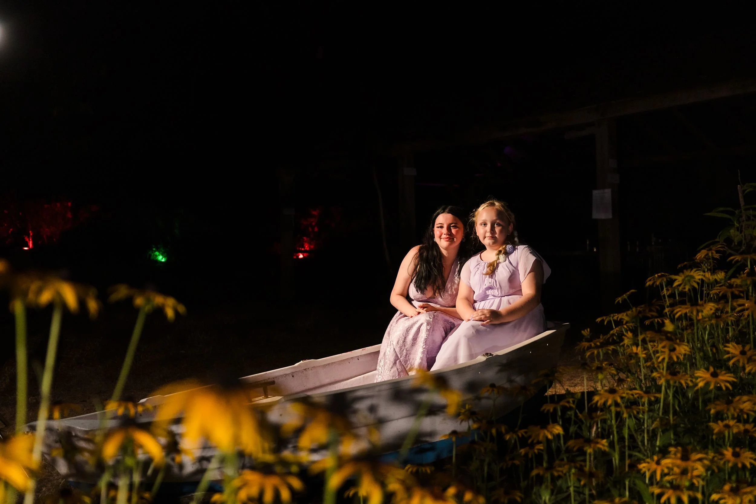 Two young wedding guests sit in the rowing boat in the walled garden at Berry Head Hotel, lit with flash after dark
