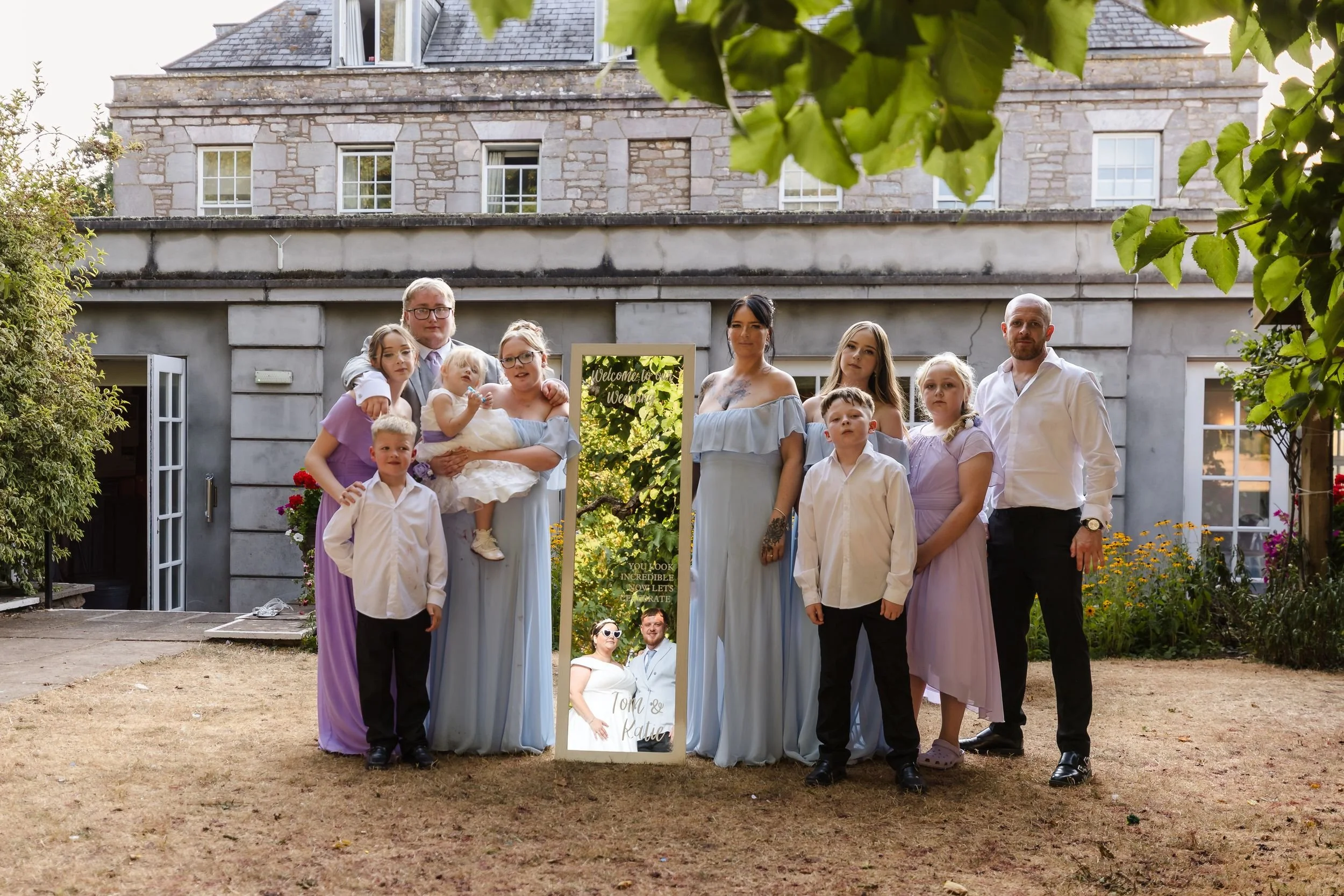 Bridal party stands around a welcome mirror in the walled garden at Berry Head Hotel, the couple reflected in the glass