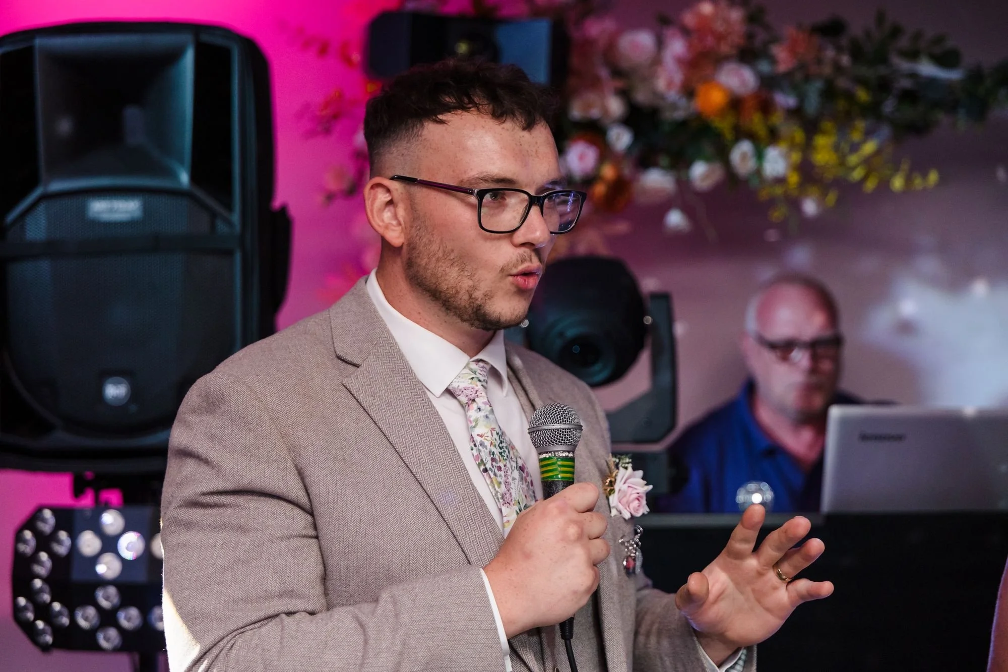 Dan speaking into a microphone in the evening room, with the DJ behind him and pink lighting on the wall