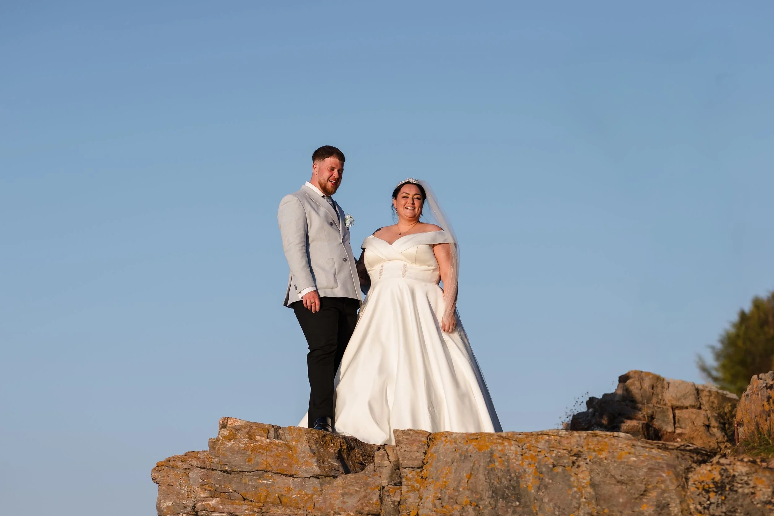 Low-angle portrait of the bride and groom laughing on the rocks at Berry Head in warm evening sun