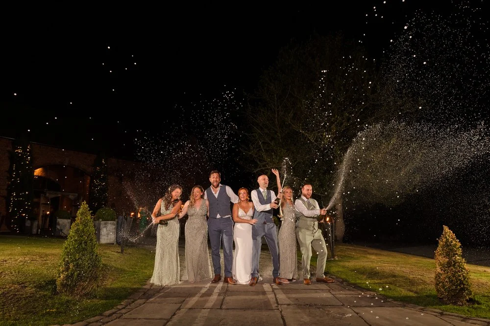 Bride, groom and wedding party spraying champagne in the grounds of Shustoke Barn at night, bridesmaids in silver sequin gowns and groomsmen in grey and sage suits celebrating with big smiles against a dark sky