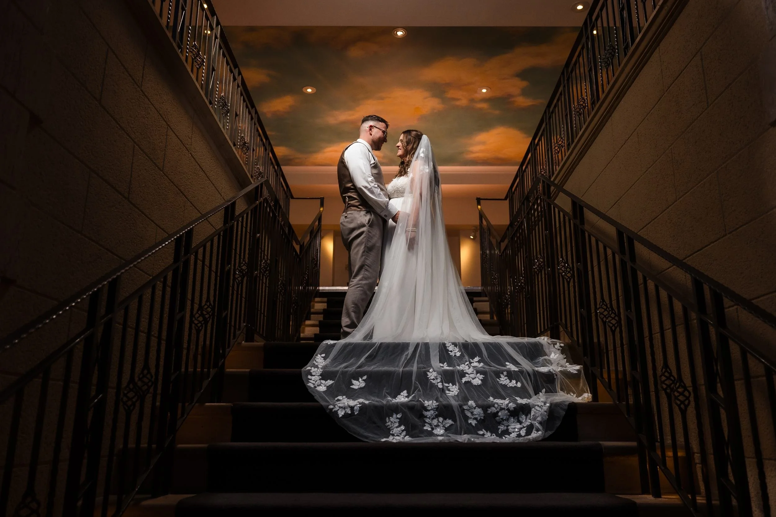 Holly and Dan facing each other on the staircase at Nailcote Hall, her veil spread down the steps, lit by warm off-camera flash