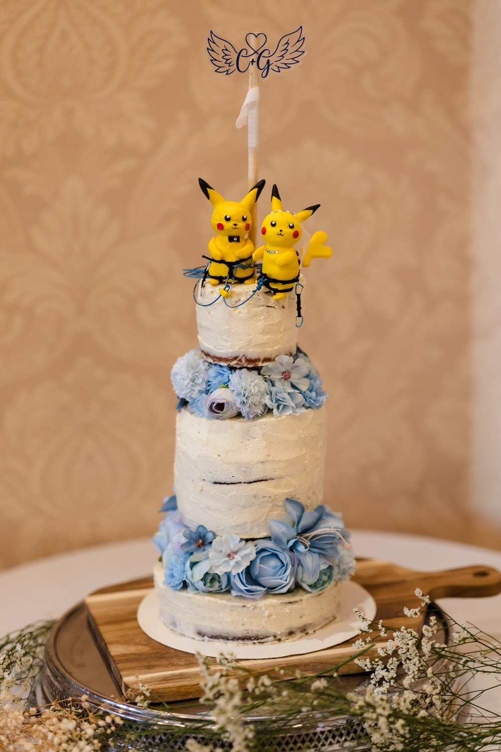 Three-tier wedding cake with blue floral details and Pikachu climbing-themed cake toppers at The Welcombe Hotel reception.