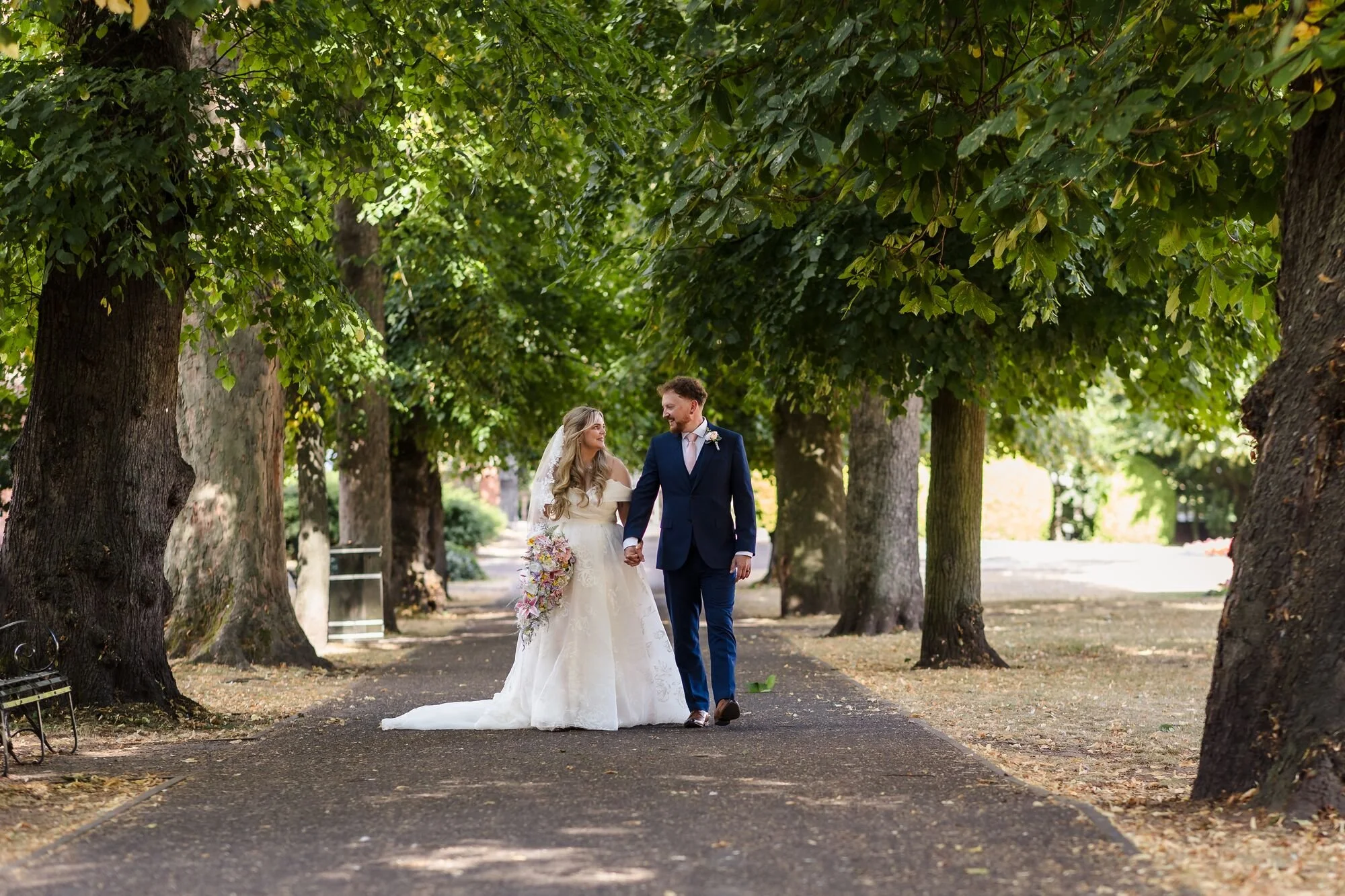 The bride and groom walking a tree-lined avenue in Museum Gardens, Lichfield, wedding photography by Once in a Lifetime Photography.