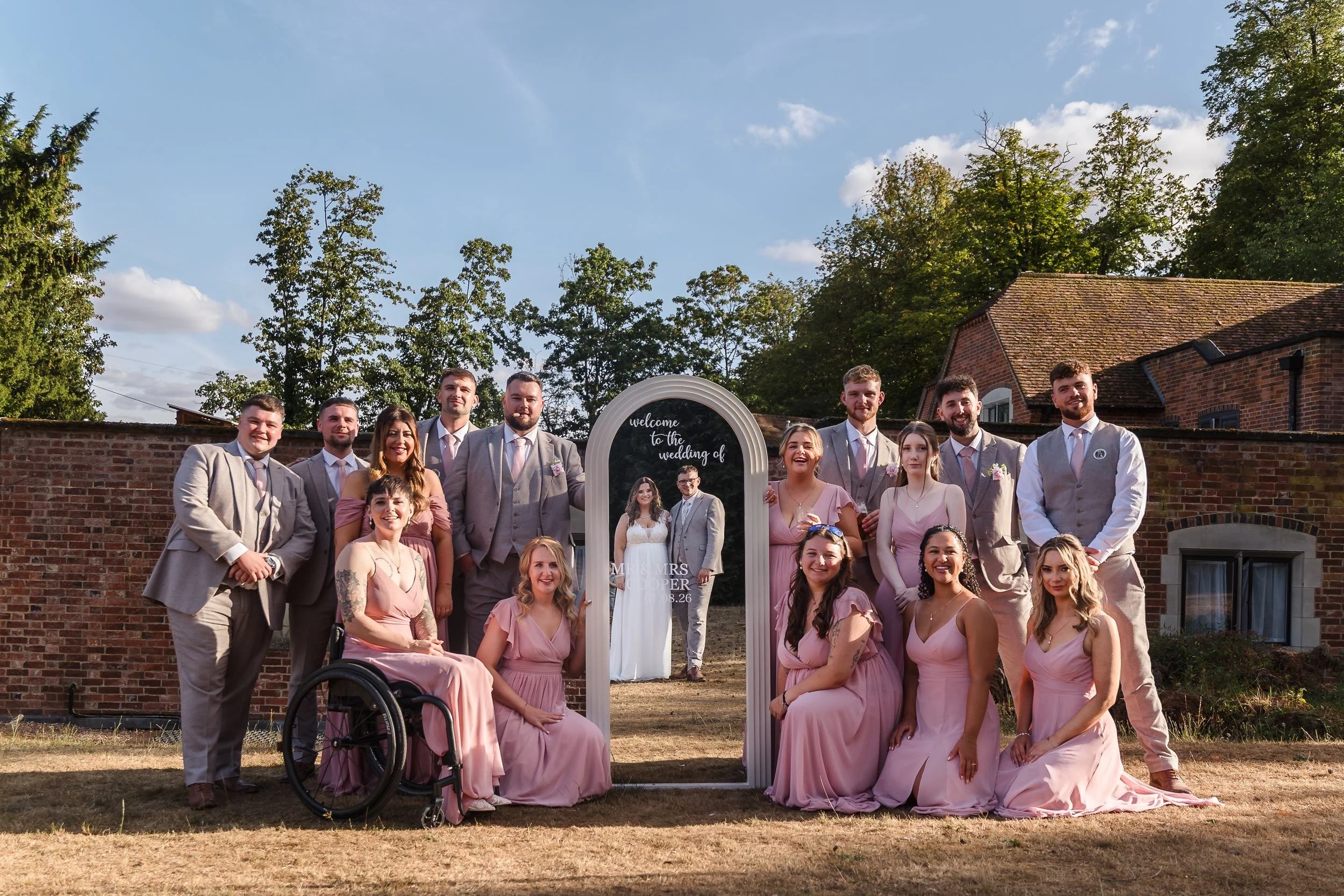 Bridesmaids and groomsmen gathered around an arched welcome mirror on the lawn, with Holly and Dan appearing as a reflection in the mirror