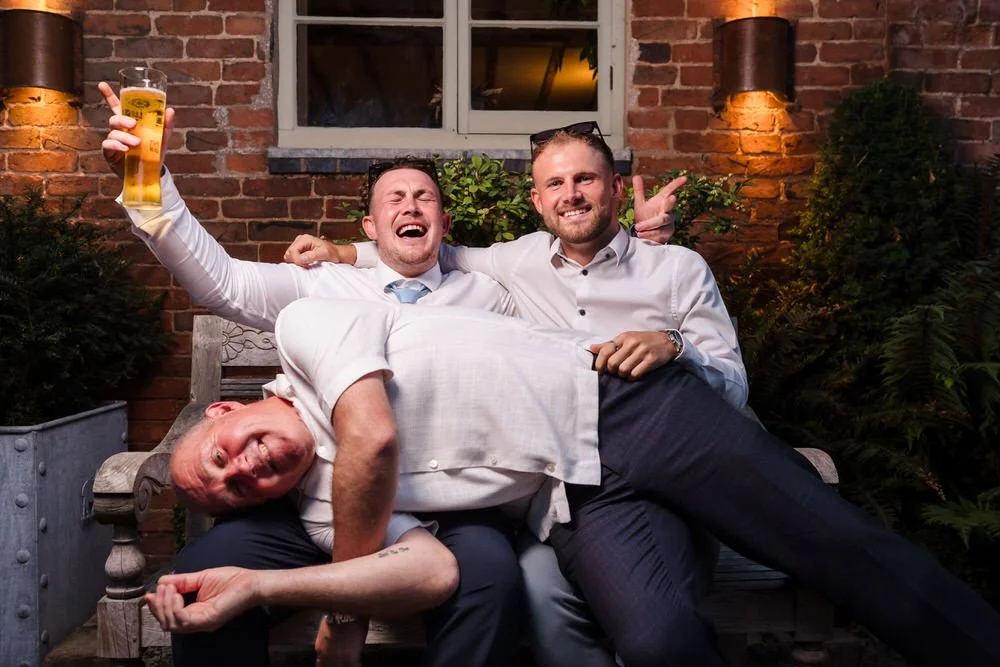 Three wedding guests larking about on a bench in the evening courtyard at Shustoke Barn, one holding a pint aloft and laughing, another lying across the bench, warm wall lighting and brick backdrop behind them