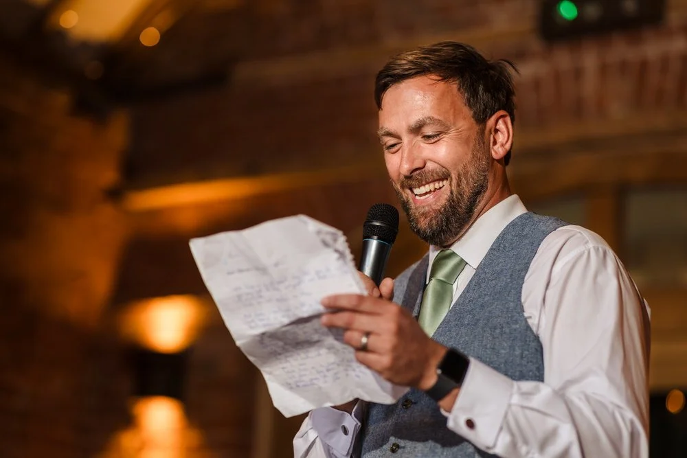 Smiling groomsman delivering a wedding speech at Shustoke Barn, holding a microphone and handwritten notes, wearing a grey waistcoat and sage green tie against the venue's warm amber-lit brick walls