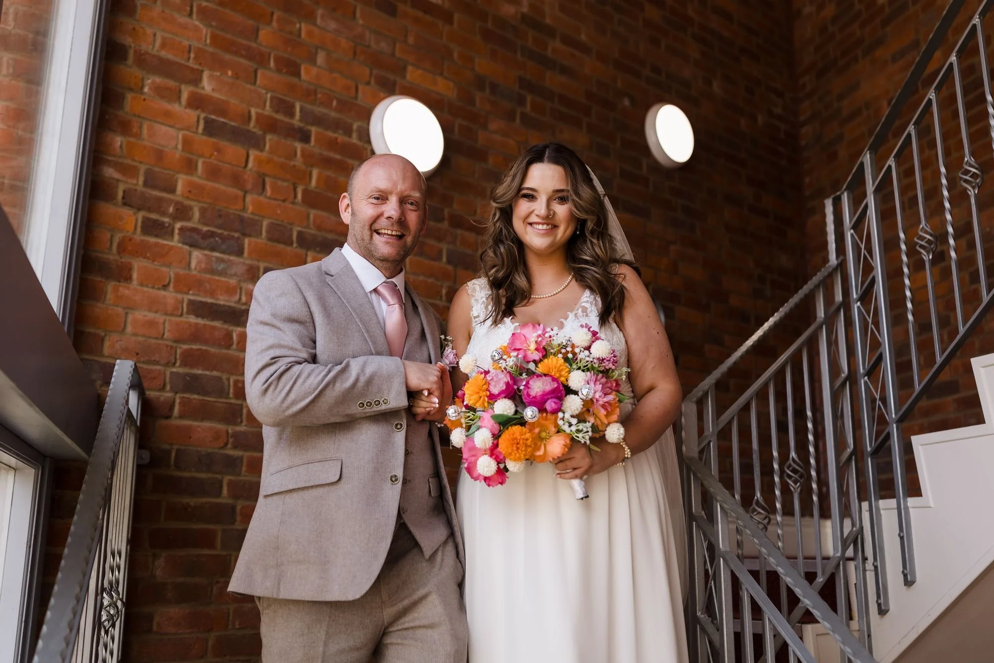 Holly holding her bright bouquet and smiling beside her dad on the staircase at Nailcote Hall before the ceremony