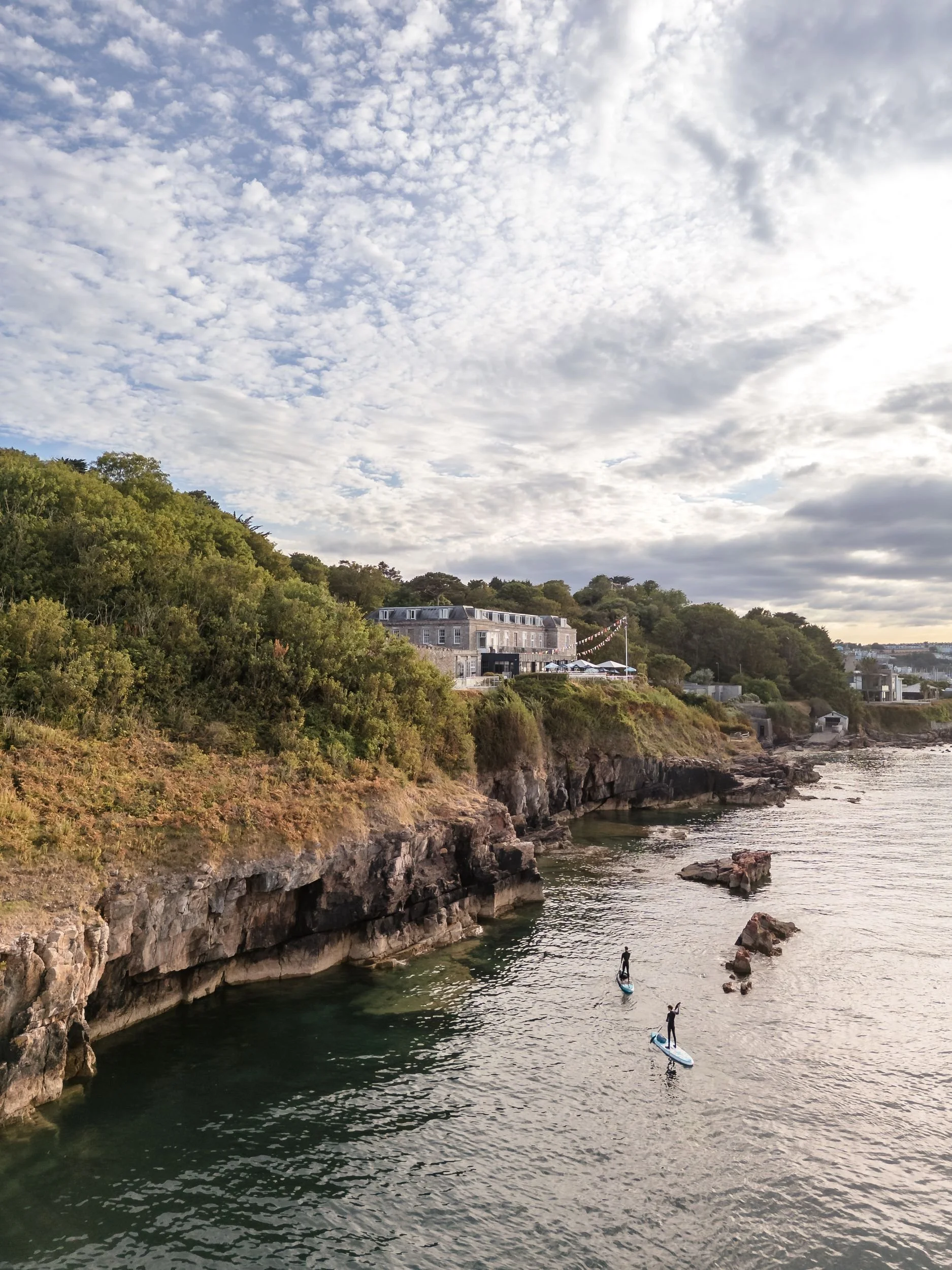 Drone view of Berry Head Hotel above the cliffs in Brixham, with paddleboarders on the water below