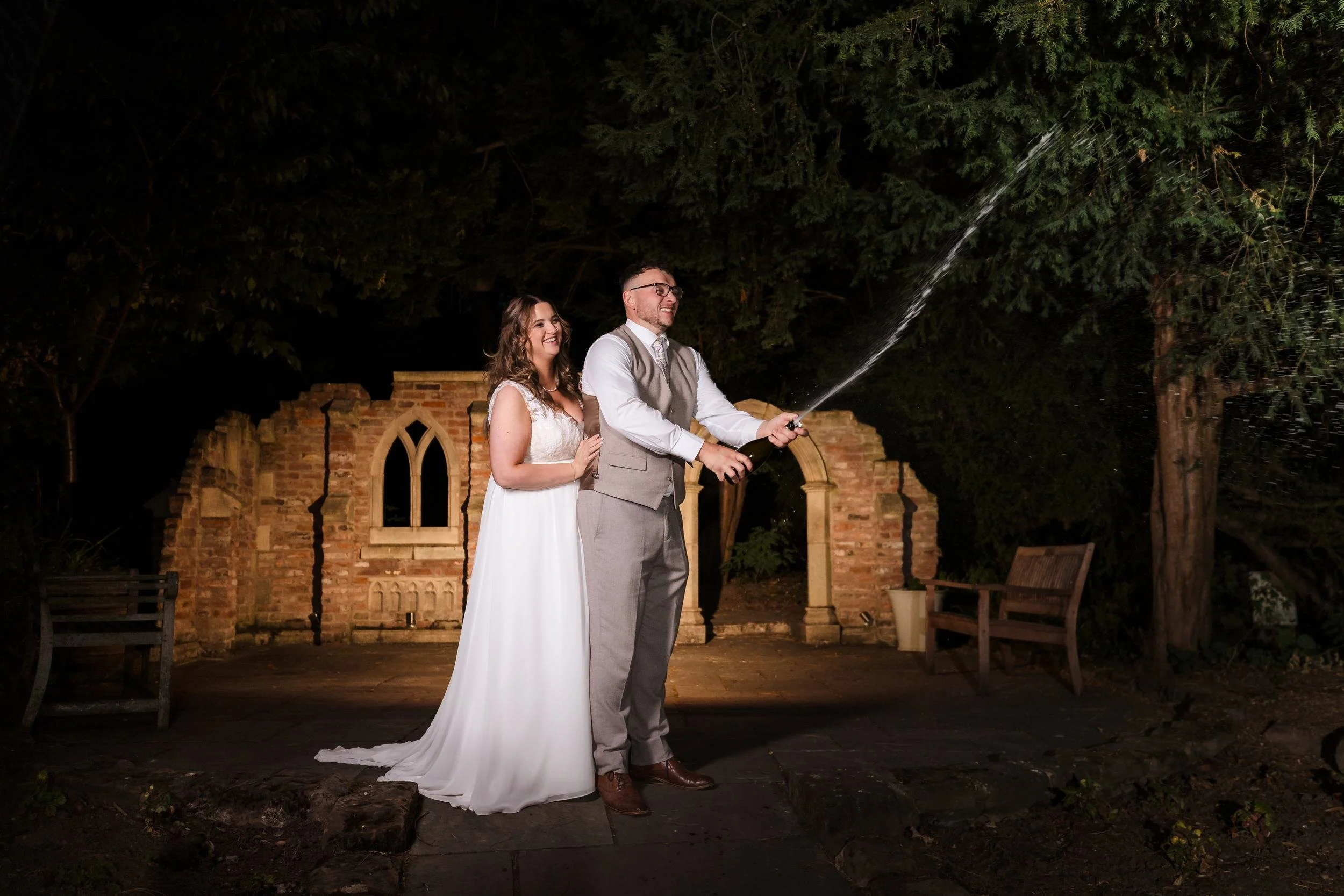 Dan spraying a bottle of champagne into the night as Holly laughs behind him, in front of the brick ruins at Nailcote Hall