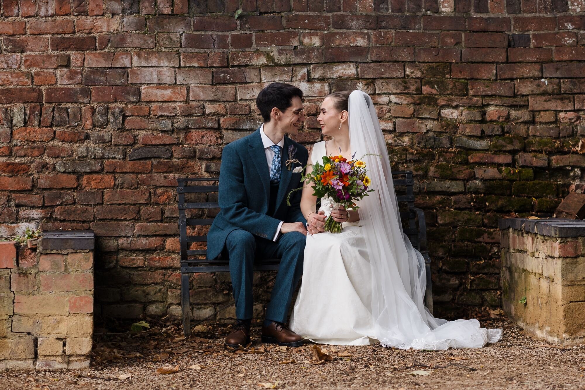 Isabelle and Chris sitting on a garden bench against a weathered brick wall at The Commandery, Worcester, by Once in a Lifetime Photography