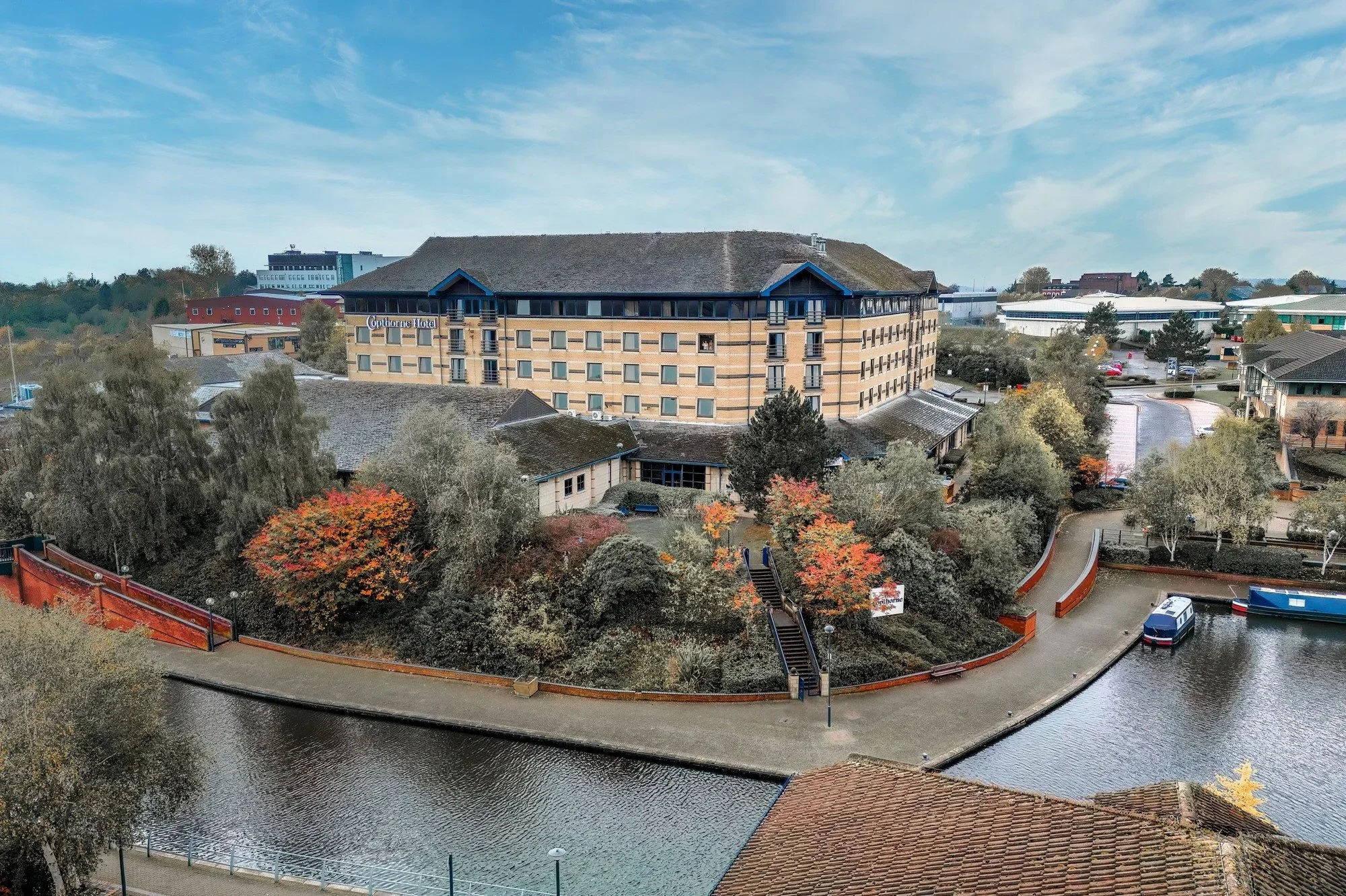 A large hotel with tan brick walls, situated above a canal with boats, surrounded by trees and smaller buildings, under a partly cloudy sky, Copthorn Hotel Merryhill.