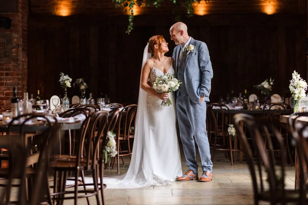 Bride and groom share an intimate moment in the warmly lit reception hall at Shustoke Barn, bride holding a white rose bouquet in a lace spaghetti strap gown, groom in a light grey three-piece suit with tan brogues