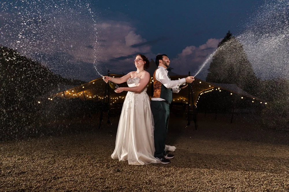Bride and groom spraying champagne at dusk in the courtyard at Huntsmill Farm, festoon lights behind.