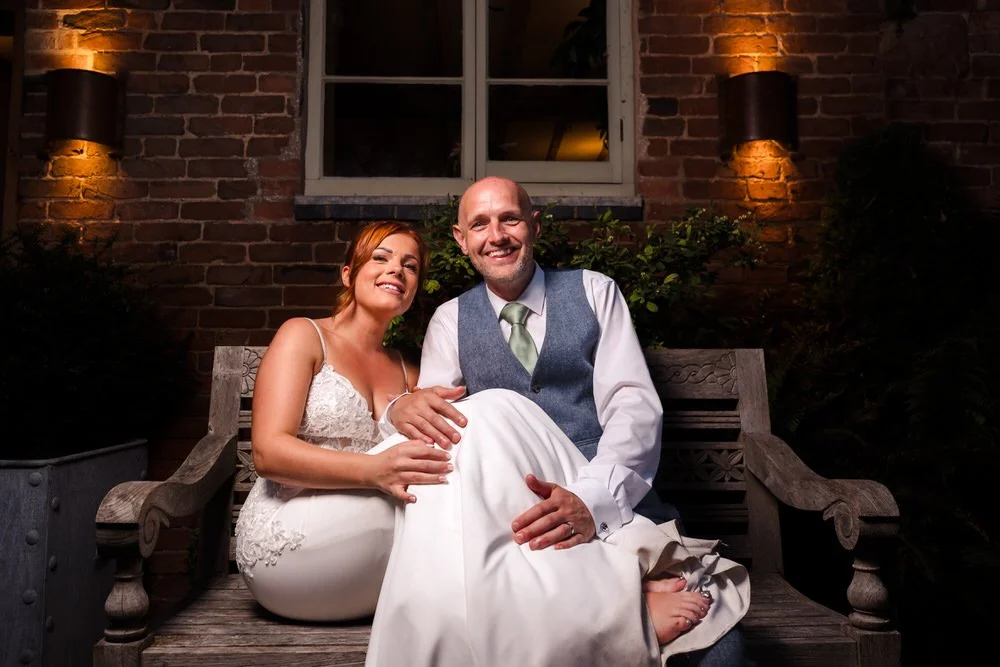 Bride and groom relaxing together on a wooden bench in the evening courtyard at Shustoke Barn, bride in lace gown leaning into groom who is wearing his grey waistcoat and sage green tie, warm wall sconces illuminating the brick backdrop behind them