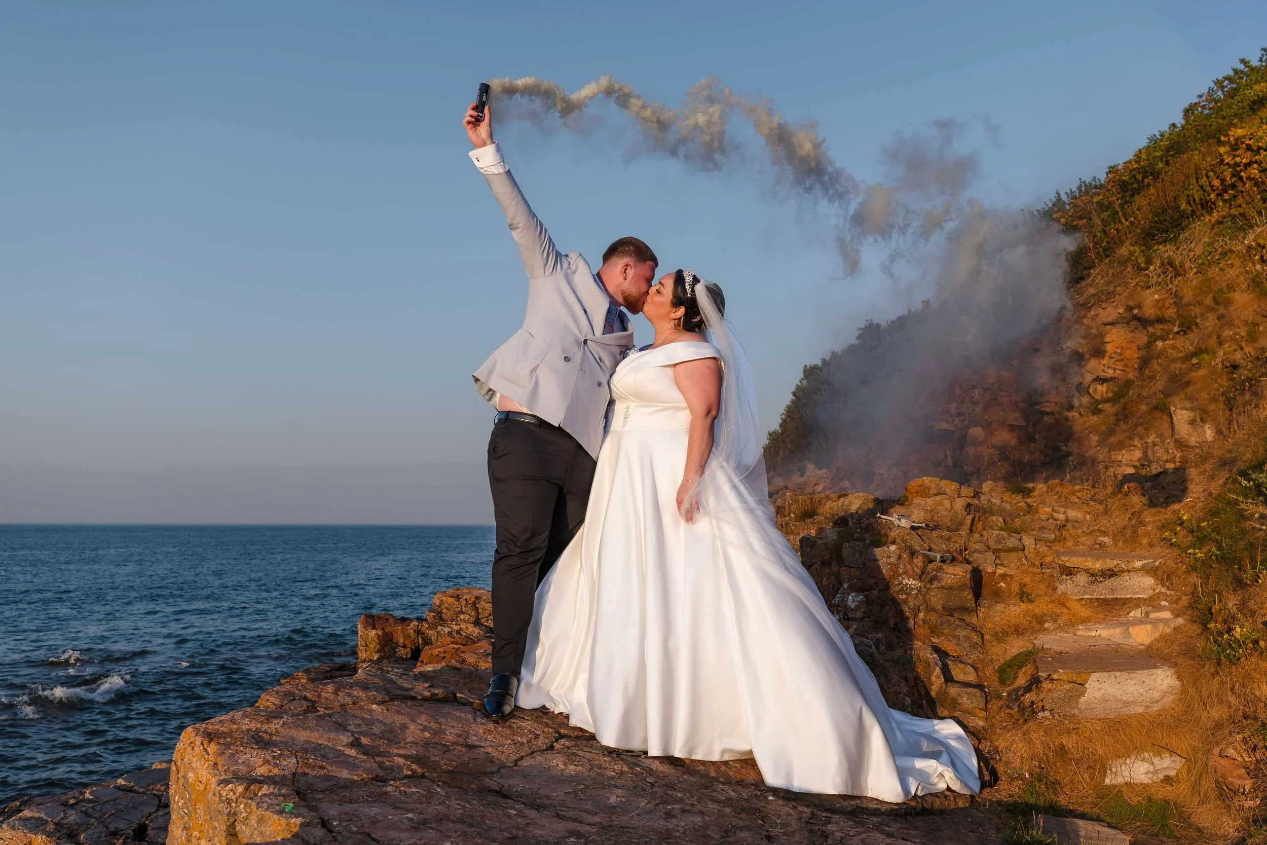 Bride and groom kiss on the rocks at Berry Head as smoke drifts across the cliff behind them