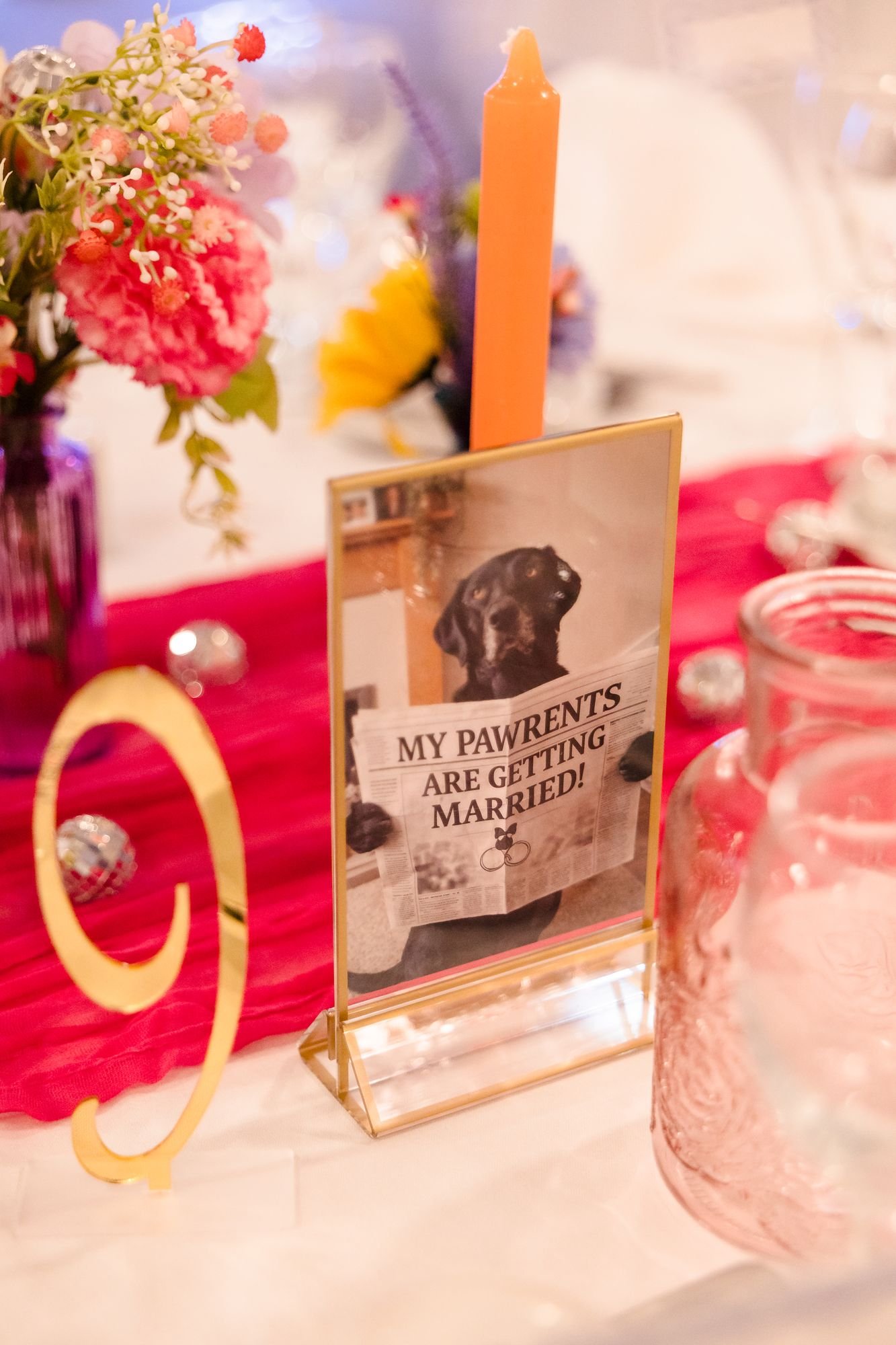 Gold-framed table sign showing the couple's dog holding a newspaper that reads My Pawrents Are Getting Married, beside an orange candle