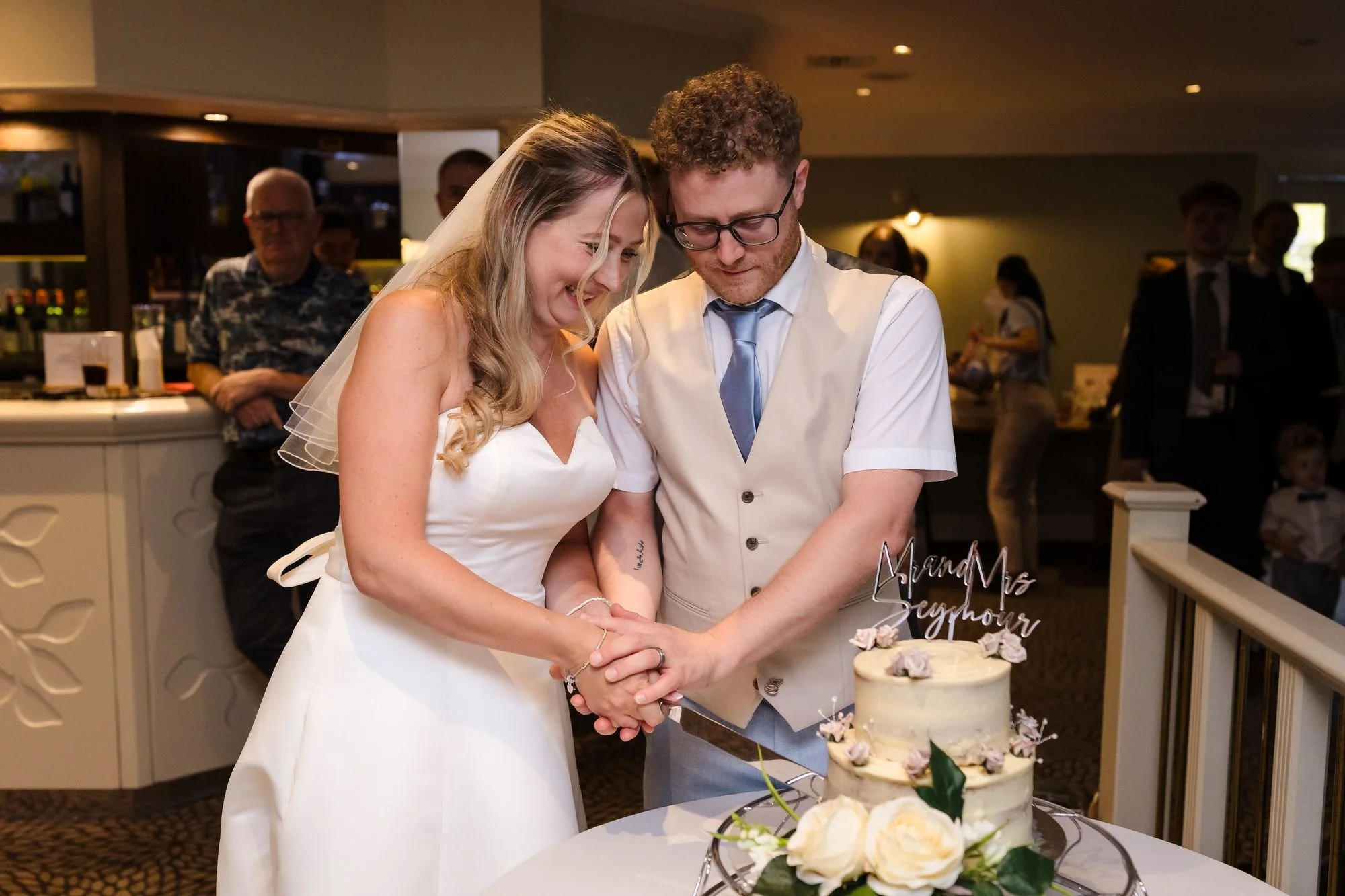 Cheryl and Josh laughing as they cut their white wedding cake with a Mr and Mrs Seymour topper