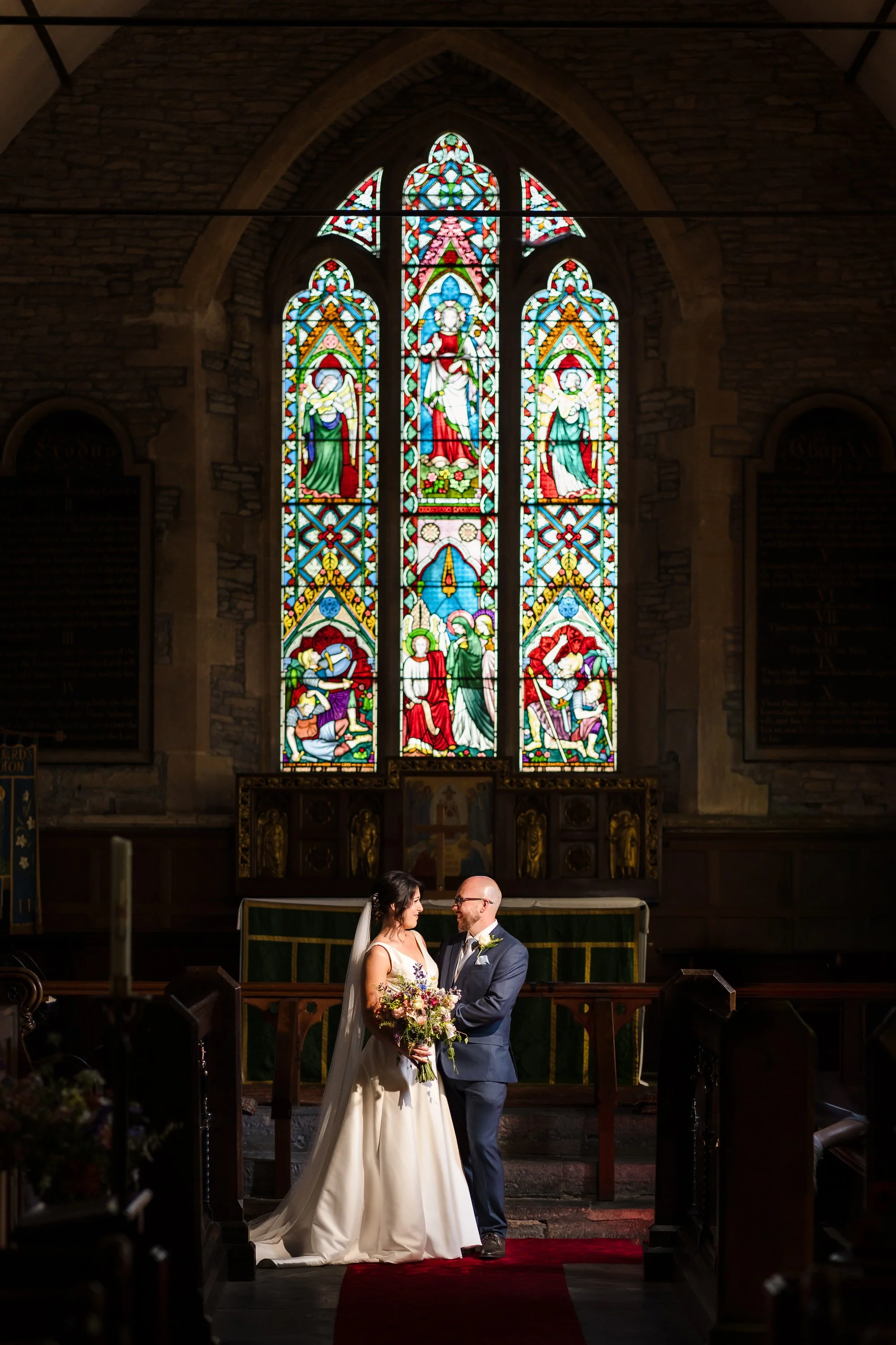 Bride and groom standing beneath a stained glass window at St Leonard's Church altar, documentary wedding photography in Worcestershire