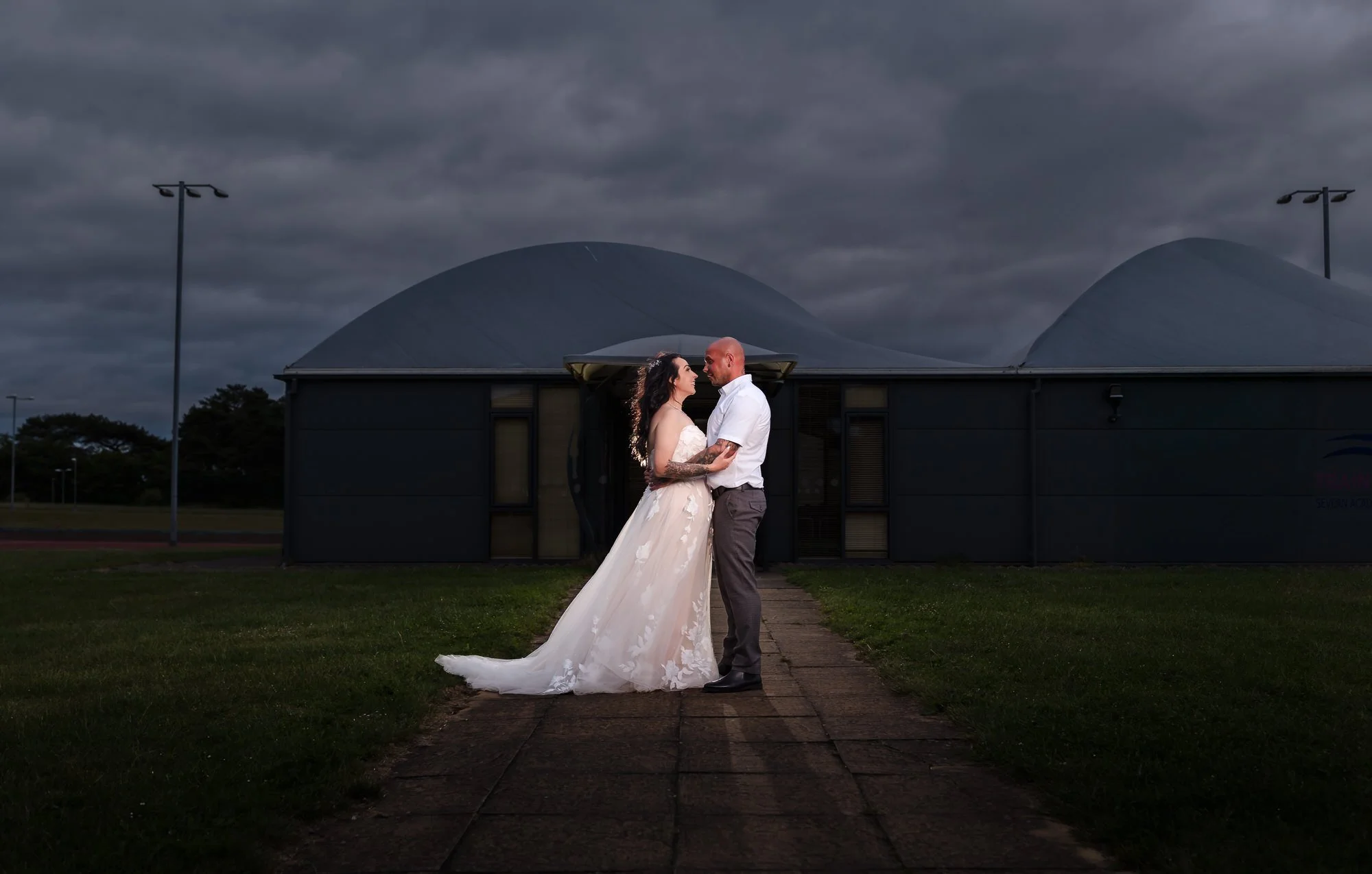 Bride and groom lit against Stourport Sports Club's clubhouse and curved roofline, with the running track behind them at dusk.