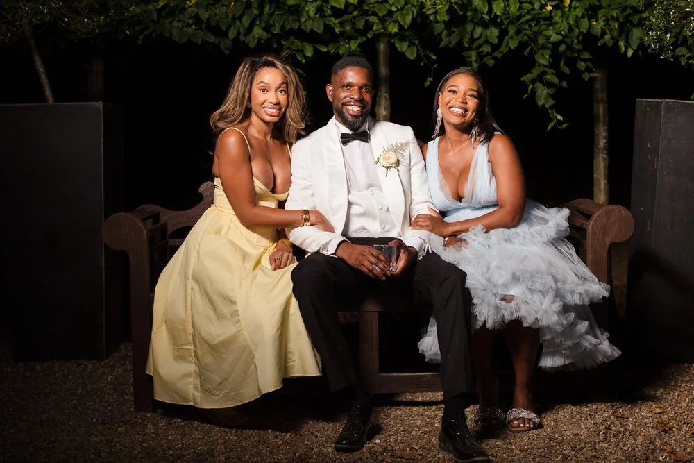 Groom seated on a bench with two guests in evening dresses, lit with off-camera flash against dark trees at Hogarths Hotel Solihull