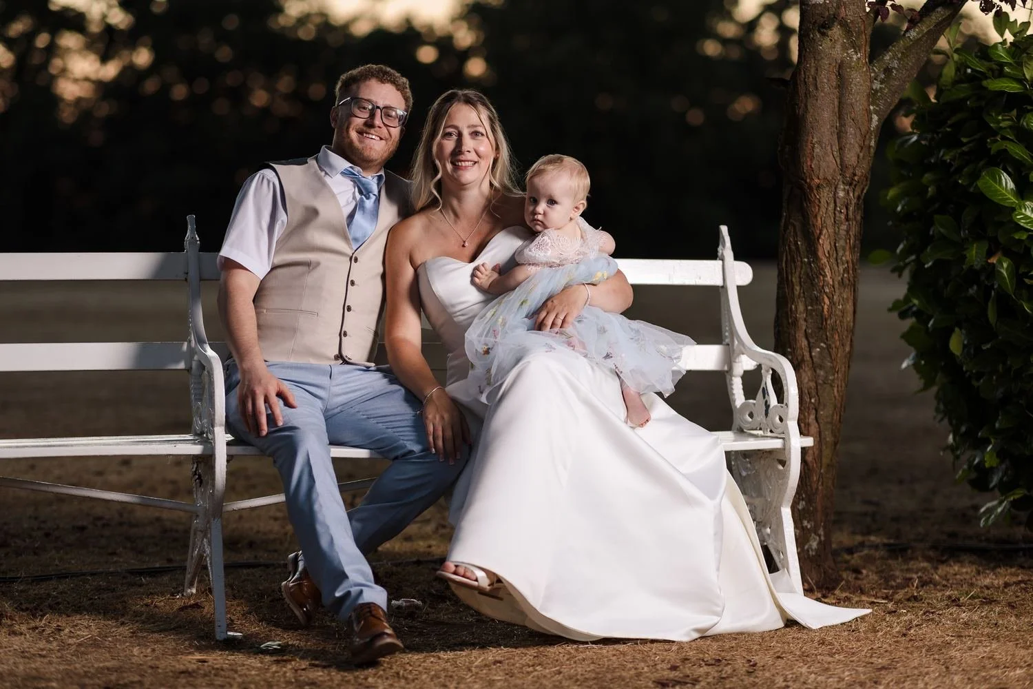 Cheryl and Josh with a baby on the white garden bench at sunset, lit with a softbox