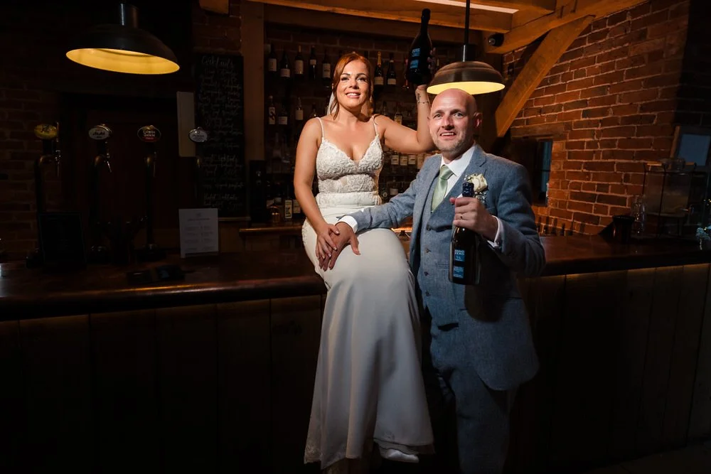 Bride sitting on the bar counter and groom holding a bottle of wine at the rustic Shustoke Barn bar, exposed brick walls and pendant lighting creating a warm atmospheric glow, bride in lace gown and groom in grey three-piece suit with sage green tie