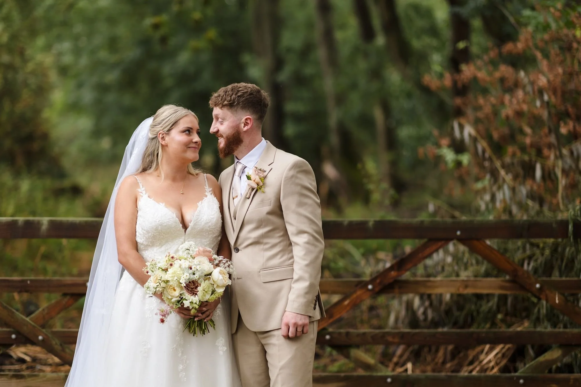 Abbie and Sean smiling at each other in front of a rustic wooden fence with woodland behind at Bennetts Willow Barn