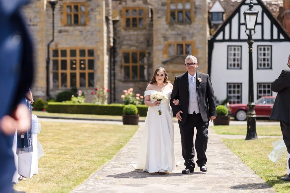 Bride walks down the outdoor aisle arm-in-arm with her father along the stone path at Karma Salford Hall.