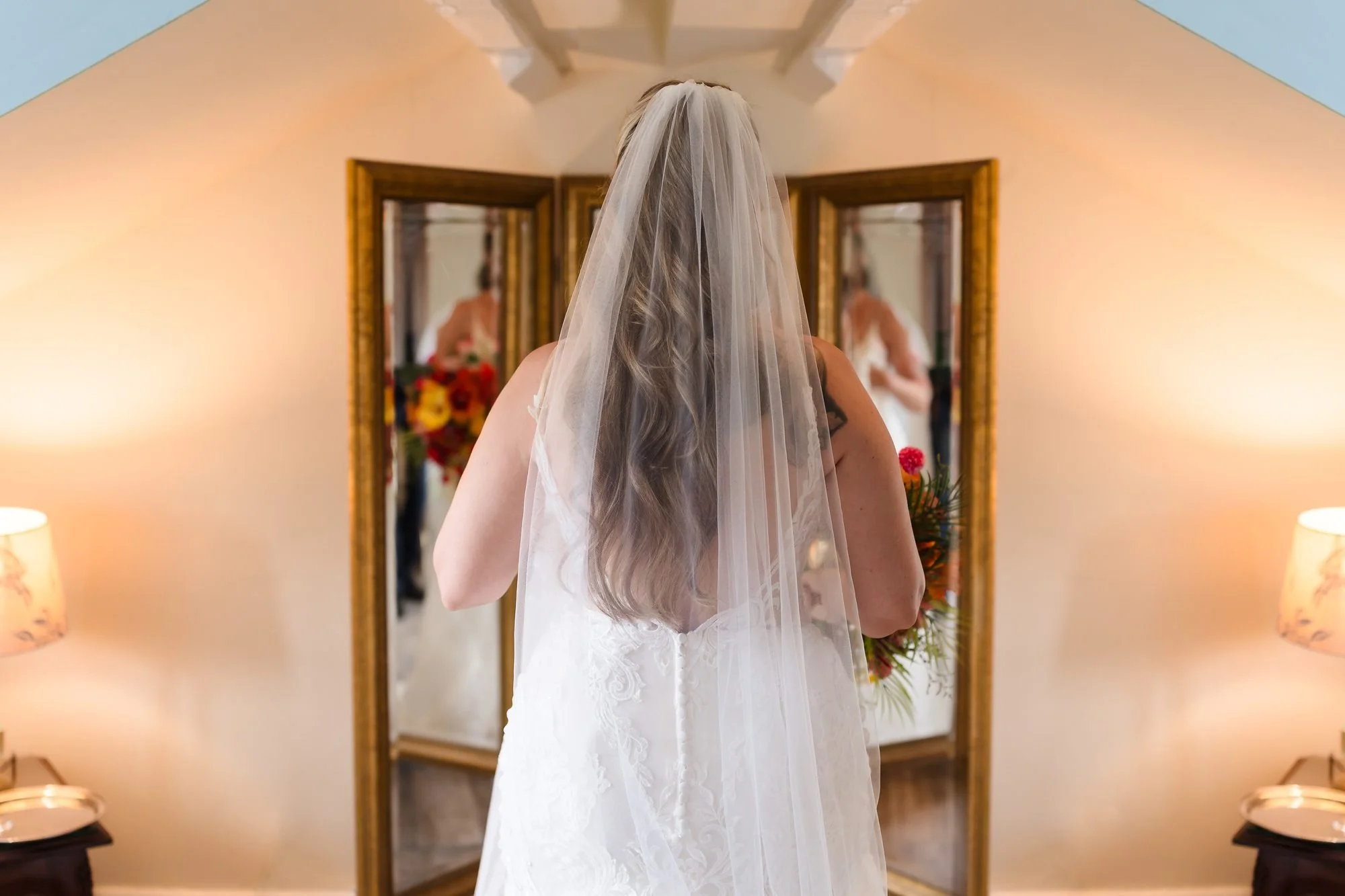 Bride Angela in her veil, seen from behind, looking into a gold mirror while getting ready upstairs at Warwick House