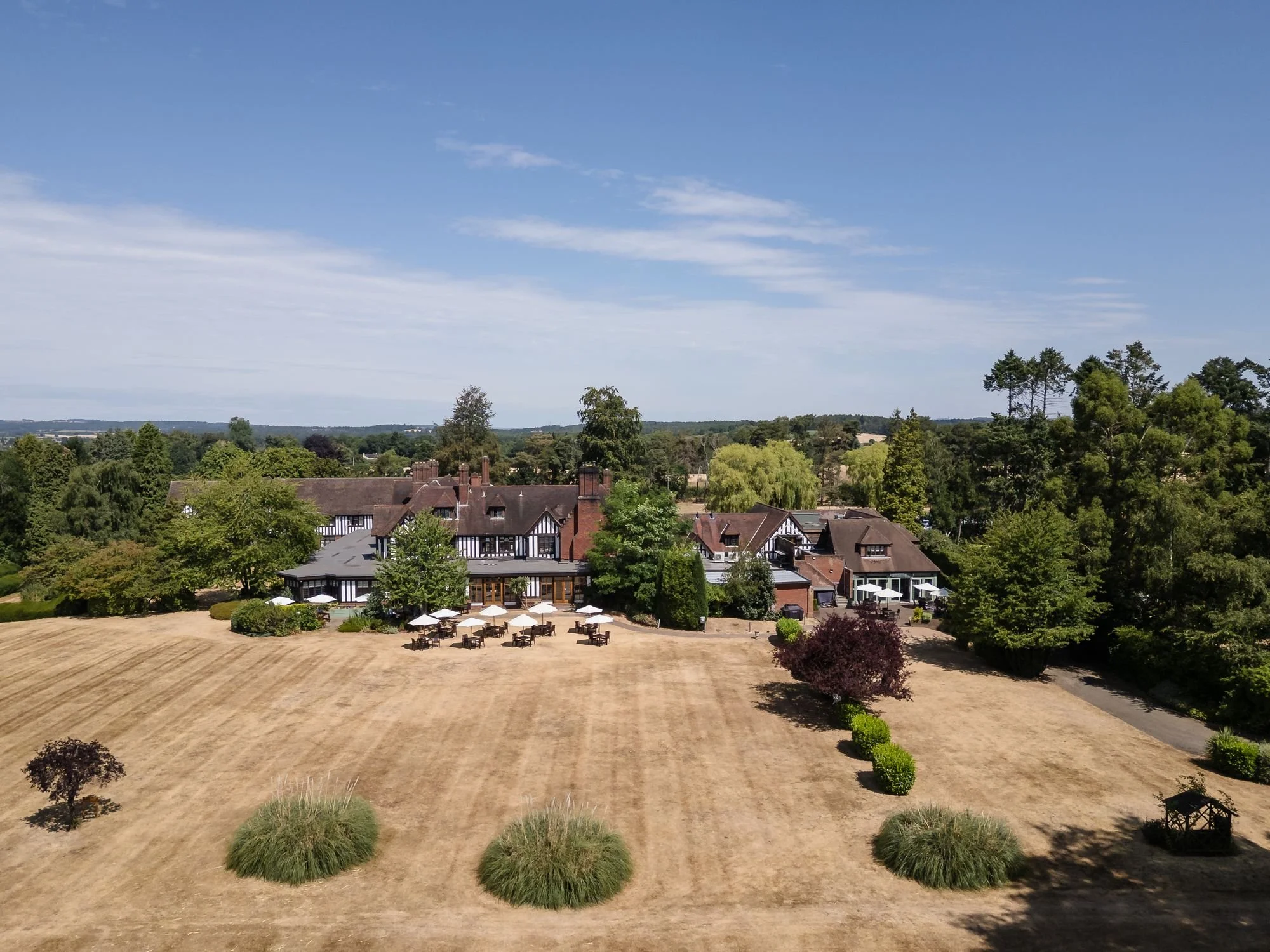Aerial view of Hogarths Stone Manor on a clear summer day, with the black-and-white timbered house, parasols on the terrace and the sweeping main lawn